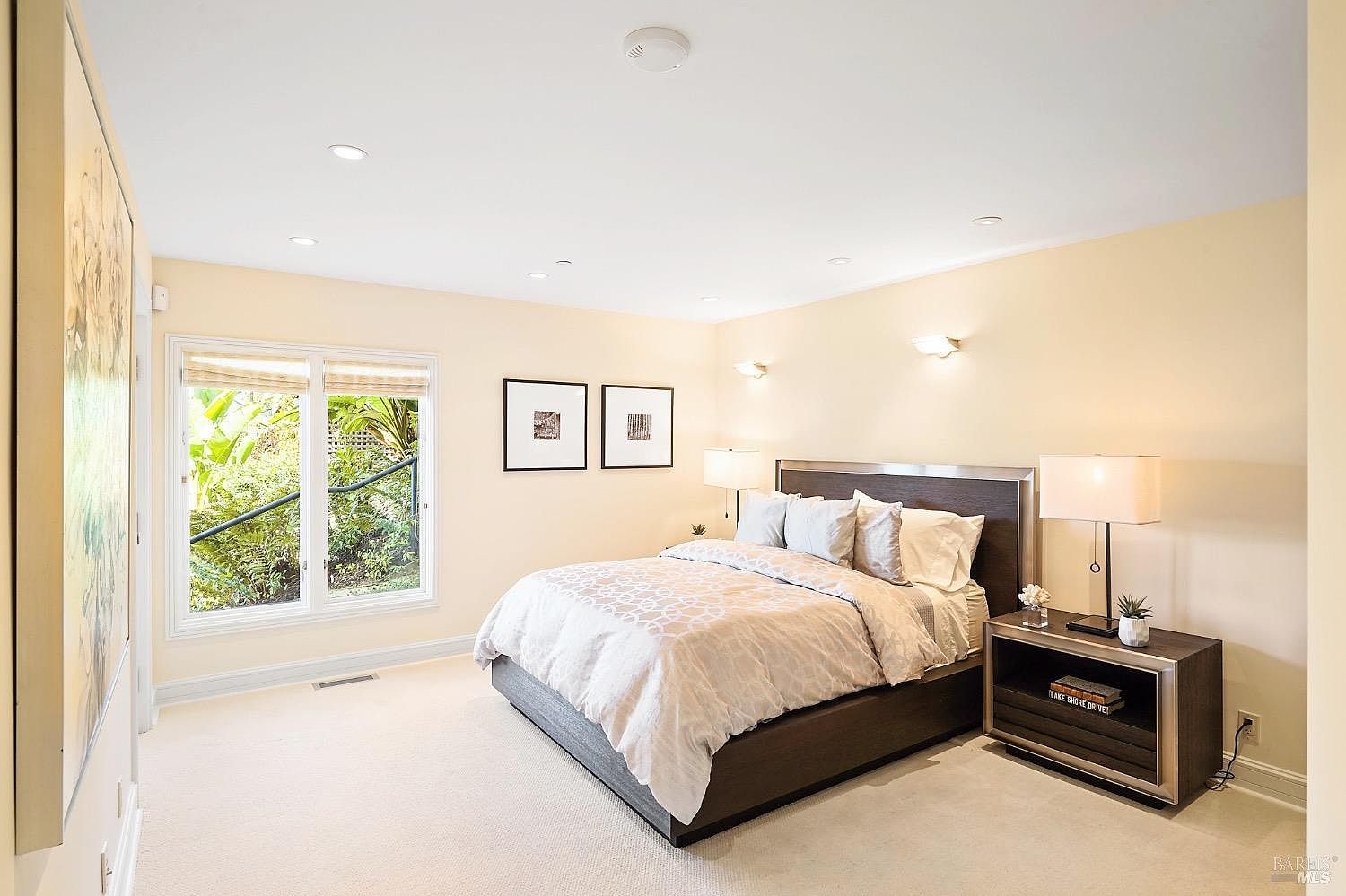 This spacious primary bedroom features a neutral color palette, plush carpeting, and a large window that provides a view of the lush greenery outside. The room is anchored by a dark-toned bed frame with a matching nightstand and is illuminated by recessed lighting and wall-mounted sconces. Two framed art pieces hang above the bed, contributing to a clean, serene, and well-appointed atmosphere.