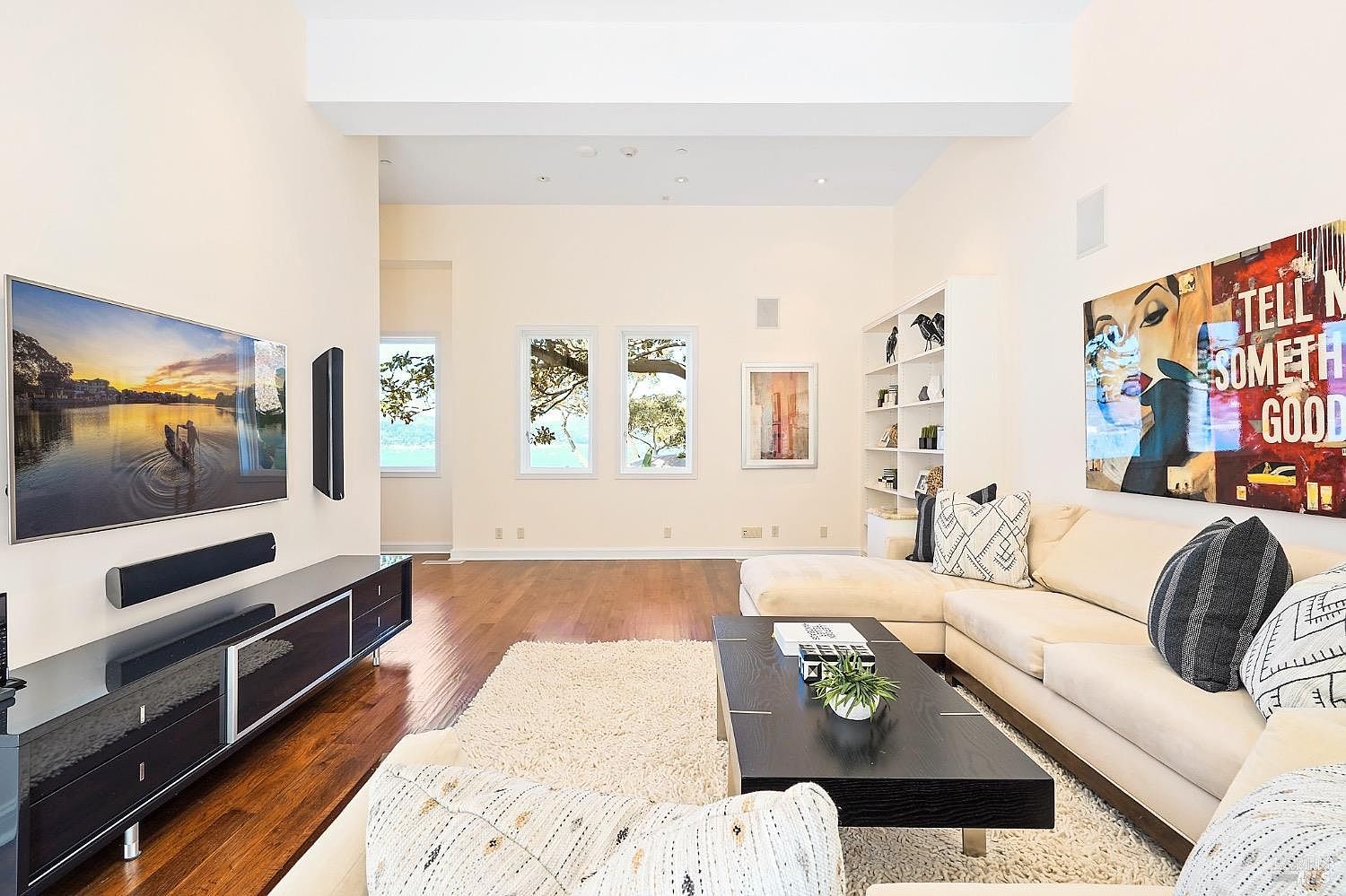 This spacious, light-filled living room features high ceilings and warm wood flooring, creating an inviting and airy atmosphere. The room is anchored by a large, cream-colored sectional sofa, a dark coffee table, and a plush area rug, while a wall-mounted television and a vibrant piece of pop-art add modern character. Large windows along the back wall provide natural light and a view of the outdoors, complemented by built-in shelving and a clean, minimalist aesthetic.