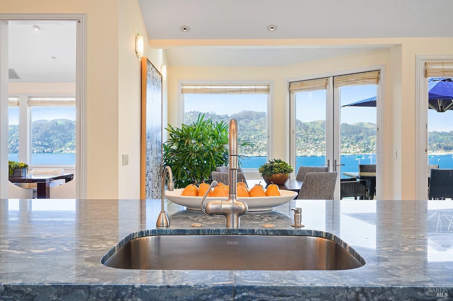 This kitchen features a prominent island with a deep, blue-toned stone countertop and a stainless steel sink, serving as the focal point of the space. Beyond the island, a dining area is visible through large glass doors that frame a stunning, expansive view of the bay and surrounding hills. The room is bright and airy, with a neutral color palette that emphasizes the connection between the indoor living space and the scenic outdoor landscape.