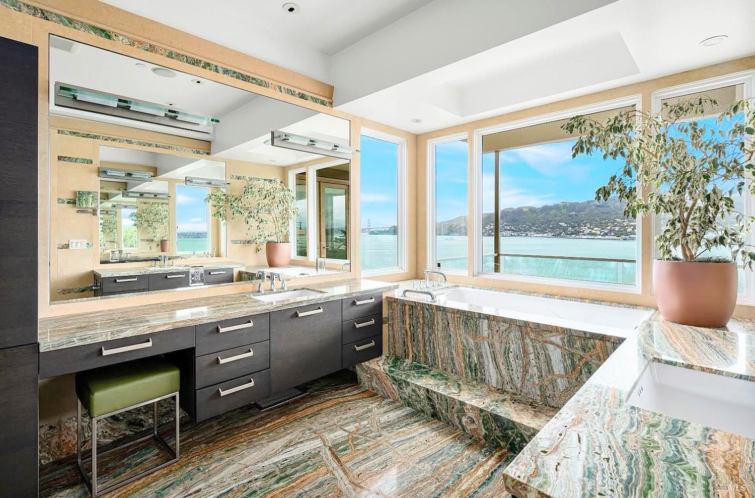 This luxurious primary bathroom features striking, heavily veined green and brown stone surfaces that cover the floor, vanity countertop, and the built-in bathtub surround. A large, wall-to-wall mirror reflects the space, while expansive windows provide panoramic water views and natural light. The dark wood cabinetry provides a sharp contrast to the vibrant stone, creating a sophisticated and high-end atmosphere.