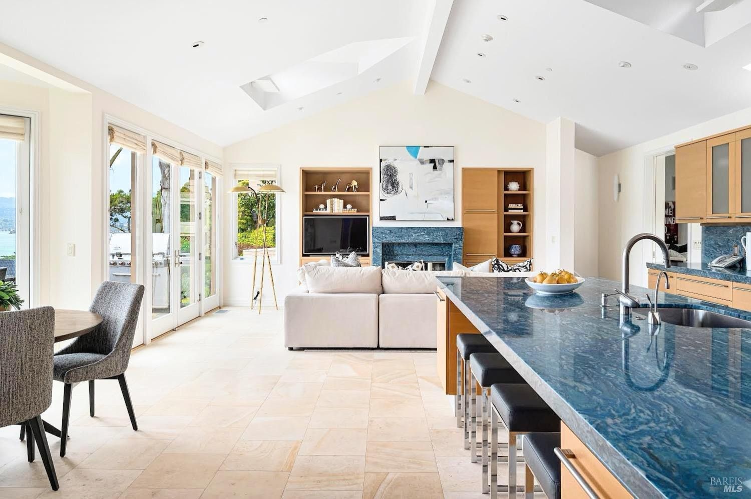This open-concept living space features a prominent kitchen island with a striking blue stone countertop and integrated bar seating. The adjacent living area is anchored by a blue stone fireplace surround and built-in shelving, all set against a backdrop of vaulted ceilings and light-toned tile flooring. Large glass doors provide a seamless transition to the outdoors, bathing the entire room in natural light and offering a view of the water.