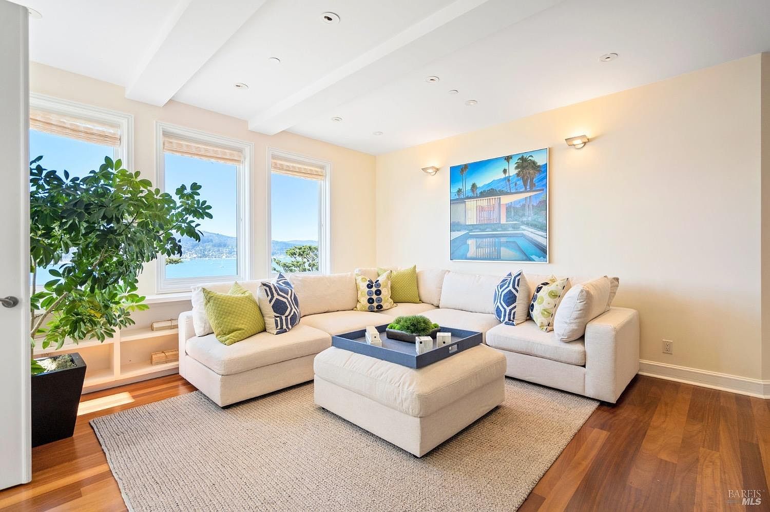This bright and airy living room features a large, light-colored sectional sofa arranged around a central ottoman with a decorative tray. The space is anchored by a textured area rug over warm-toned wood flooring and is illuminated by natural light streaming through three large windows that offer a scenic water view. A piece of vibrant wall art adds a pop of color to the neutral walls, creating a welcoming and relaxed atmosphere.