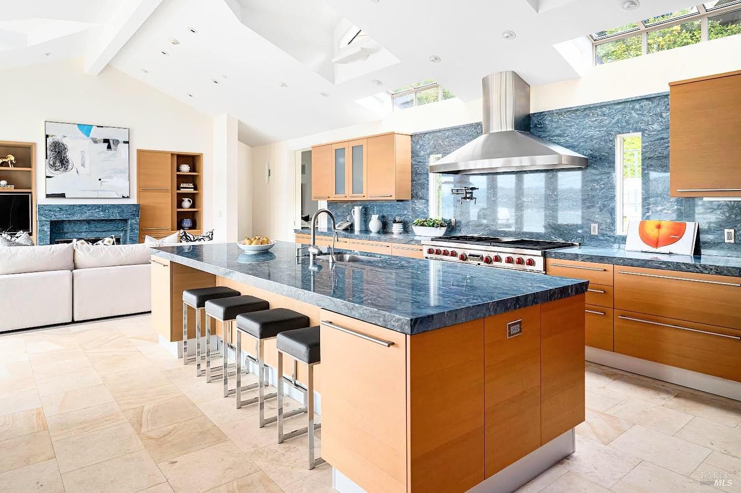 This spacious, open-concept kitchen features a large central island with a blue stone countertop and four bar stools, serving as the focal point of the room. The space is characterized by light wood cabinetry, a professional-grade stainless steel range with a matching hood, and a striking blue stone backsplash that extends across the wall. The room is bathed in natural light from multiple skylights and large windows, creating an airy atmosphere that flows seamlessly into the adjacent living area.