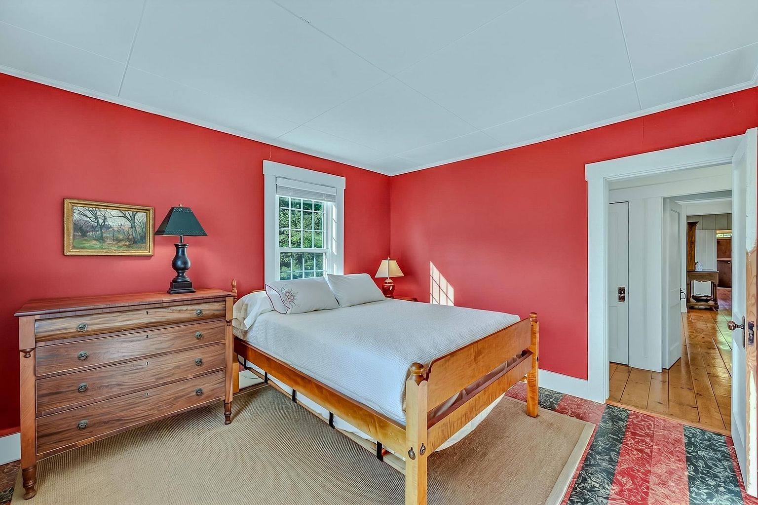 This bedroom features bold red walls contrasted by white trim and a light-colored ceiling. A wooden bed frame with white bedding sits centered in the room, flanked by a wooden dresser on the left and a small bedside lamp. The perspective looks into the room from the doorway, revealing a glimpse of a hallway with wooden flooring beyond.