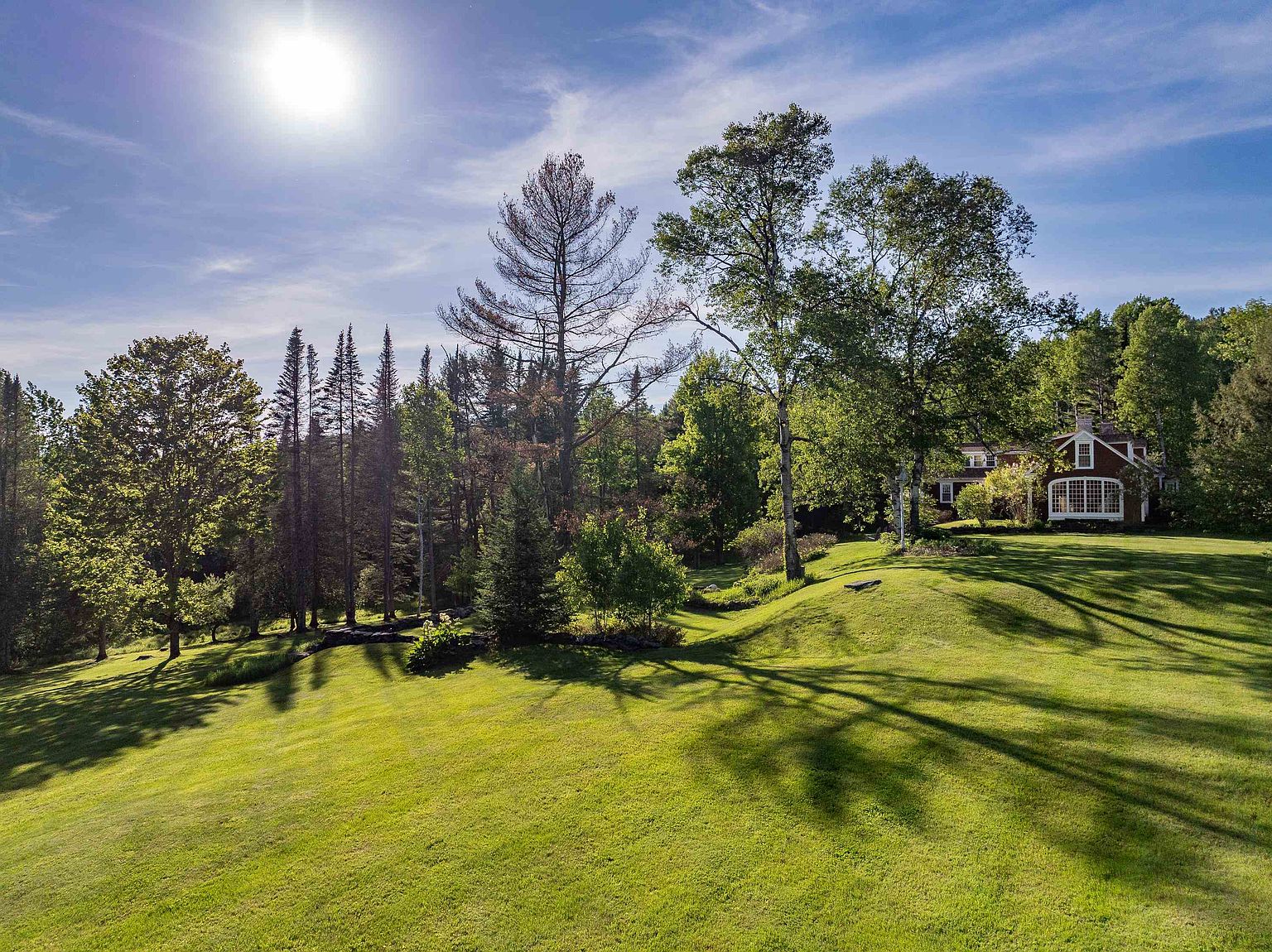 This wide-angle shot captures a sprawling, lush green lawn bathed in bright, natural sunlight under a clear blue sky. A red-sided house with a prominent multi-pane window is nestled among mature trees and dense forest foliage in the background. The perspective is low and expansive, emphasizing the vastness of the property and the serene, park-like setting.
