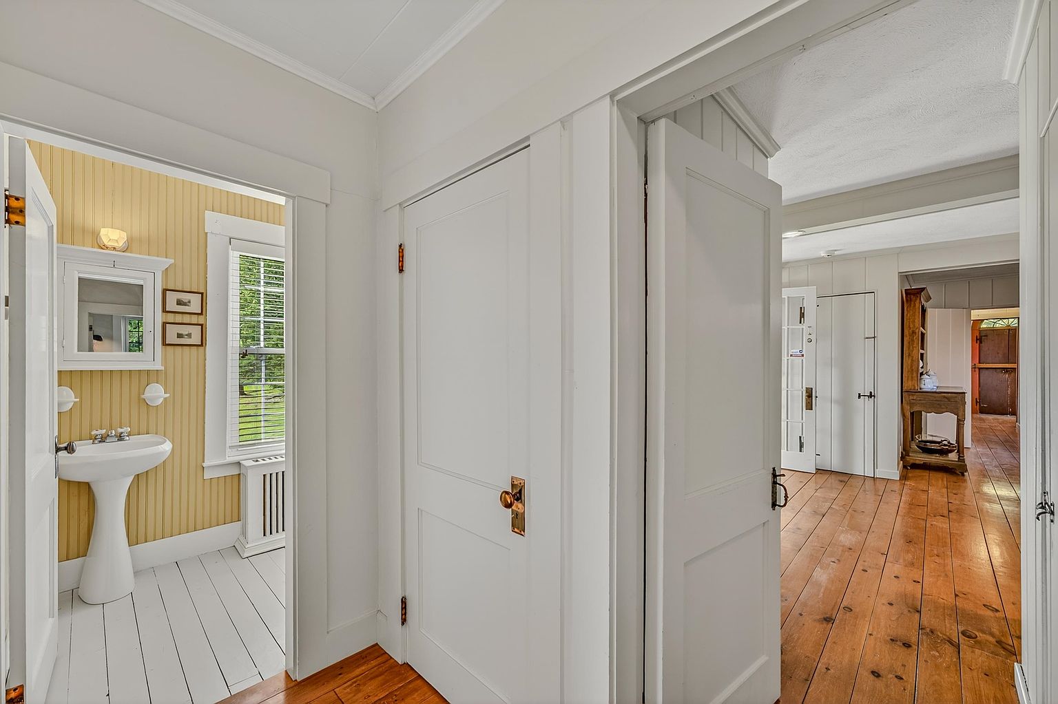 This interior view captures a transition space connecting a bathroom with yellow beadboard walls to a wider hallway with warm-toned wood flooring. A central white door with a brass knob serves as a focal point, flanked by an open doorway leading to the bathroom and a view into an adjacent room featuring a wooden hutch. The perspective emphasizes the home's traditional architectural character, highlighting the contrast between the painted white trim and the natural wood floors.