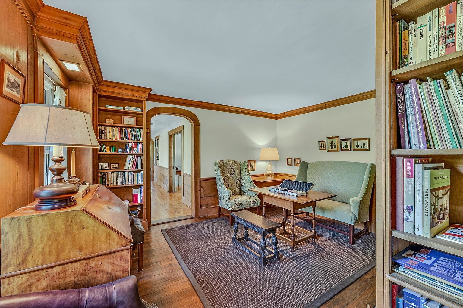 This cozy, traditional living room features warm wood-paneled walls, built-in bookshelves, and hardwood flooring. The space is furnished with a patterned armchair, a light-colored sofa, and a wooden coffee table, all centered on a textured area rug. A wide, arched doorway leads to a hallway, creating an inviting and classic atmosphere.