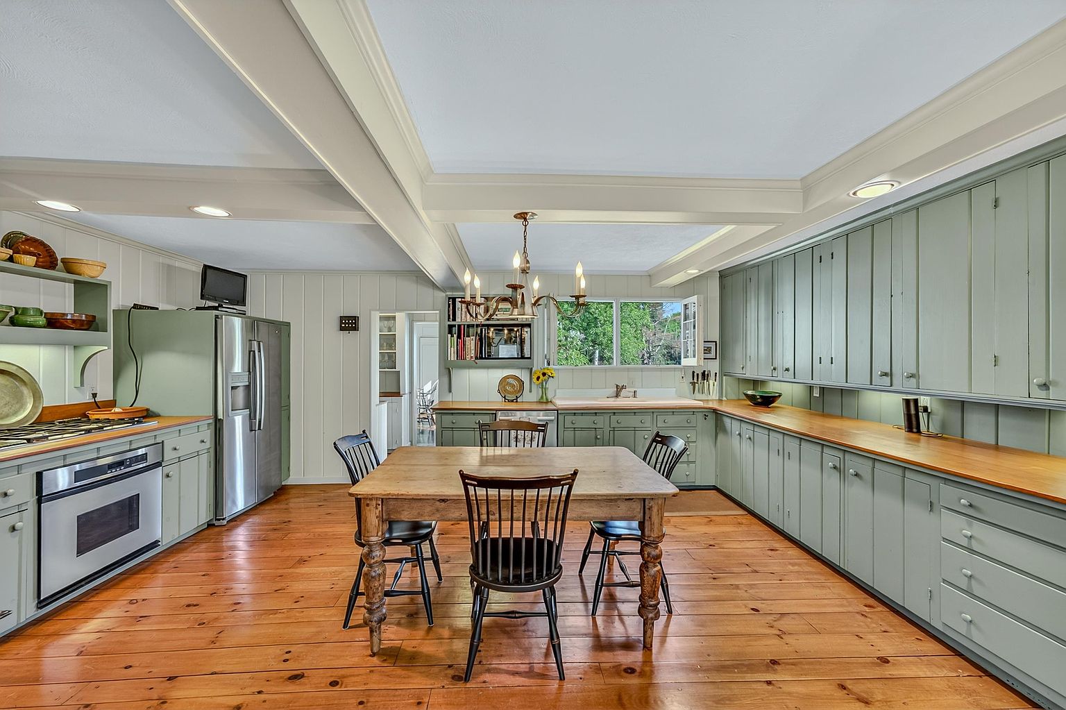 This spacious, rustic-inspired kitchen features sage green cabinetry paired with warm wood countertops and wide-plank wood flooring. A central wooden dining table serves as a focal point under a classic chandelier, while the room is framed by exposed ceiling beams and a long run of cabinetry that provides extensive storage. The perspective is a wide, symmetrical shot capturing the entire kitchen layout, including the stainless steel refrigerator and built-in oven.