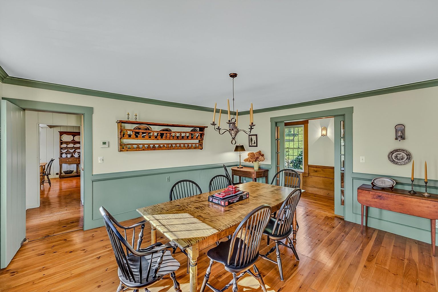 This traditional dining room features warm wood flooring and a classic color palette with sage green wainscoting. A large wooden dining table sits beneath a multi-armed chandelier, while a decorative wall-mounted plate rack adds a touch of rustic charm. The room flows into adjacent spaces through wide doorways, creating an open and inviting atmosphere.