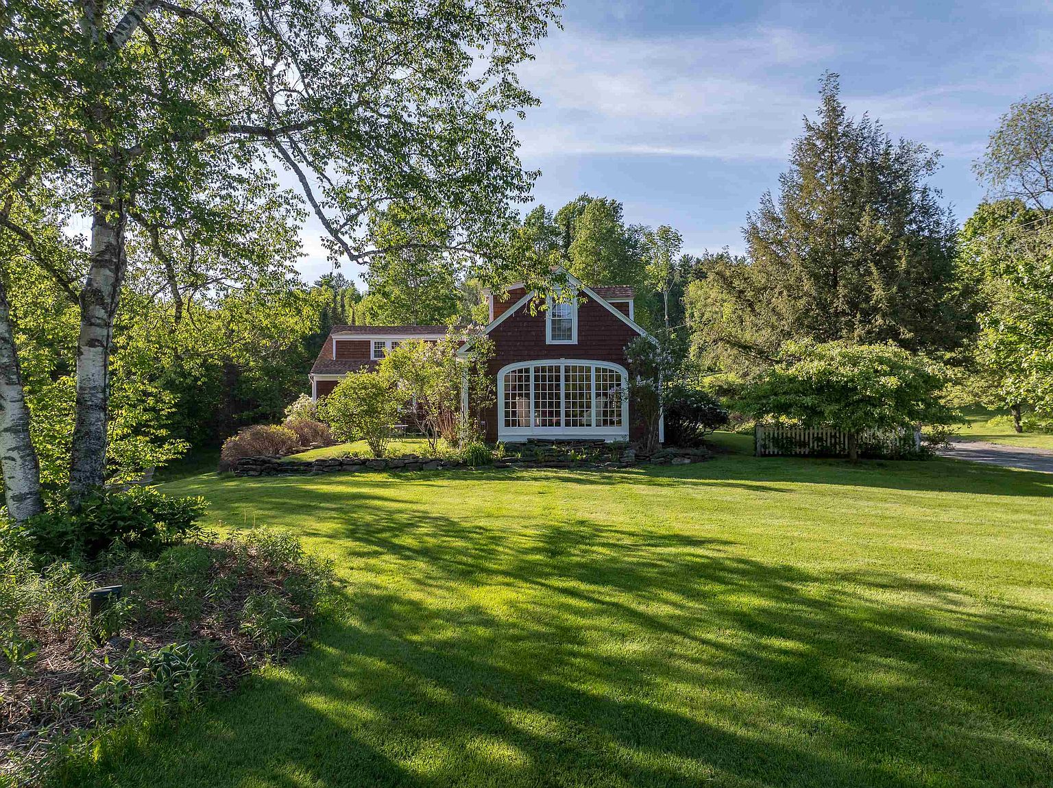 This charming, shingled home is nestled in a lush, green landscape, featuring a prominent, large multi-pane window that serves as a focal point on the front facade. The property is surrounded by mature trees and a well-manicured lawn, with a stone retaining wall adding texture to the garden beds. The perspective is a wide, eye-level shot that captures the serene, park-like setting of the residence.