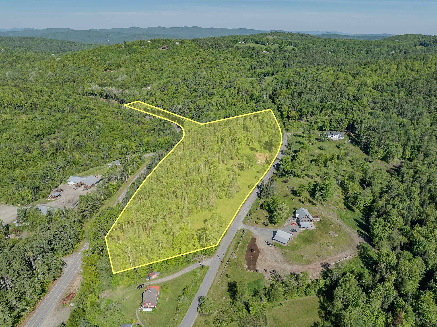This high-angle aerial view showcases a large, wooded plot of land highlighted by a yellow boundary outline. The property is situated in a rural, hilly landscape surrounded by dense forest and nearby residential homes. A paved road runs along the edge of the property, providing clear access to the site.