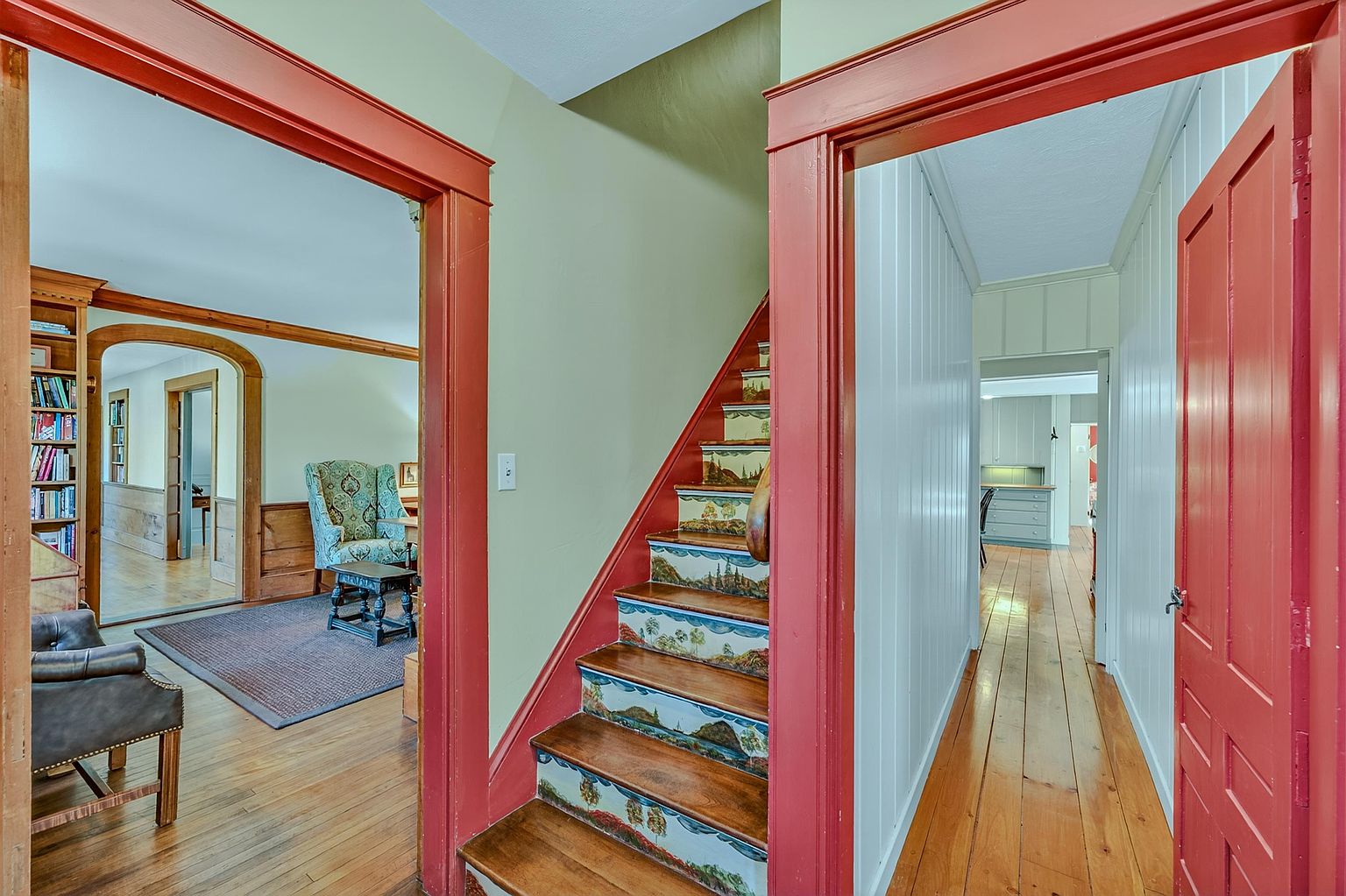 This interior view captures a central hallway featuring a staircase with decorative, landscape-painted risers and a vibrant red-painted trim. To the left, an arched doorway leads into a living area with a bookshelf and a patterned armchair, while a straight hallway to the right leads toward a kitchen space. The home showcases a blend of traditional architectural details, including light wood flooring and paneled walls, creating a warm and character-filled atmosphere.