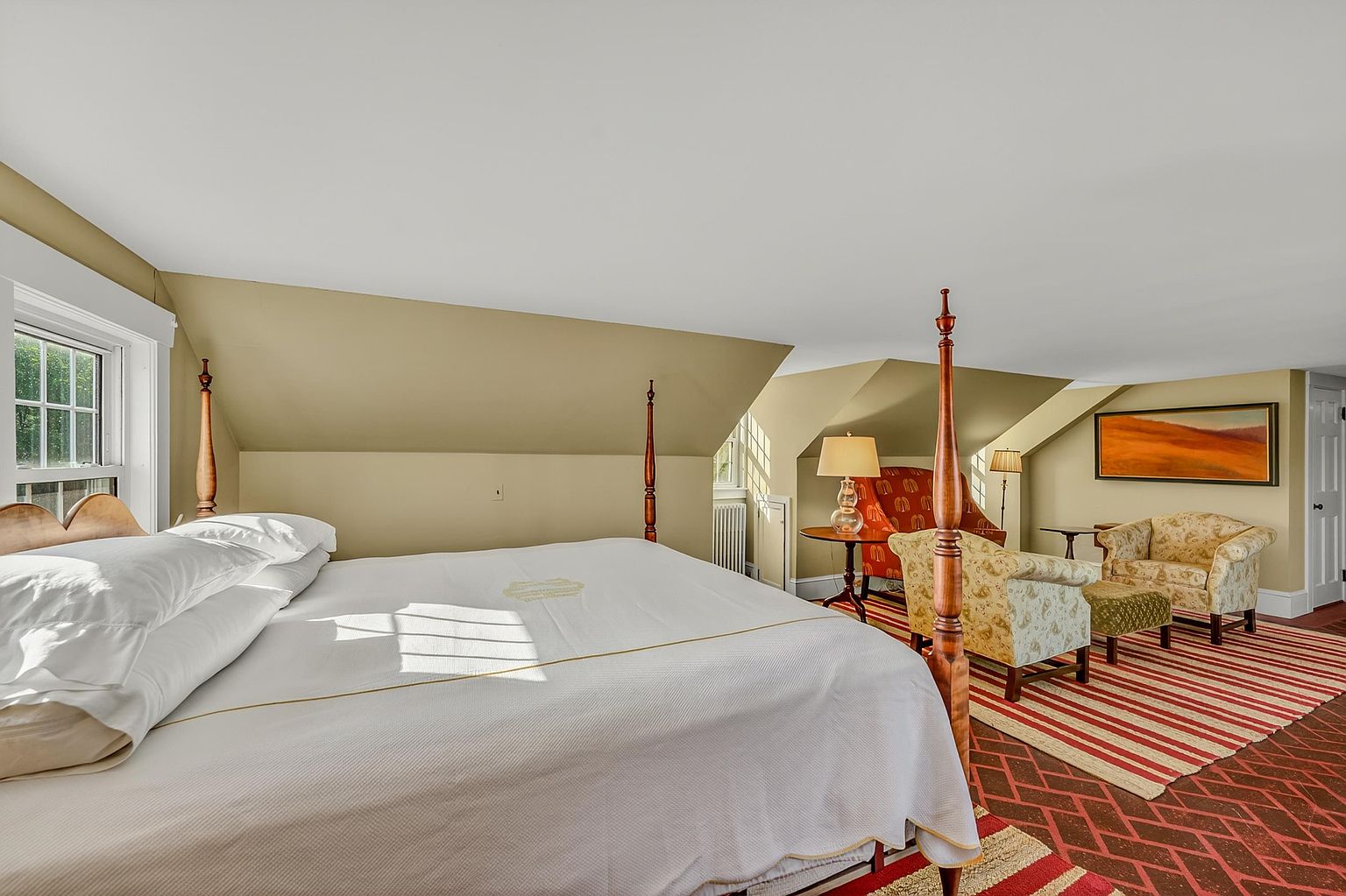 This spacious bedroom features a classic four-poster bed as the central focal point, set against a backdrop of sloped ceilings and warm-toned walls. A cozy seating area with patterned armchairs and a matching ottoman is positioned near a window, creating a comfortable reading nook. The room is finished with a red and white striped area rug over a patterned floor, offering a traditional and inviting atmosphere.