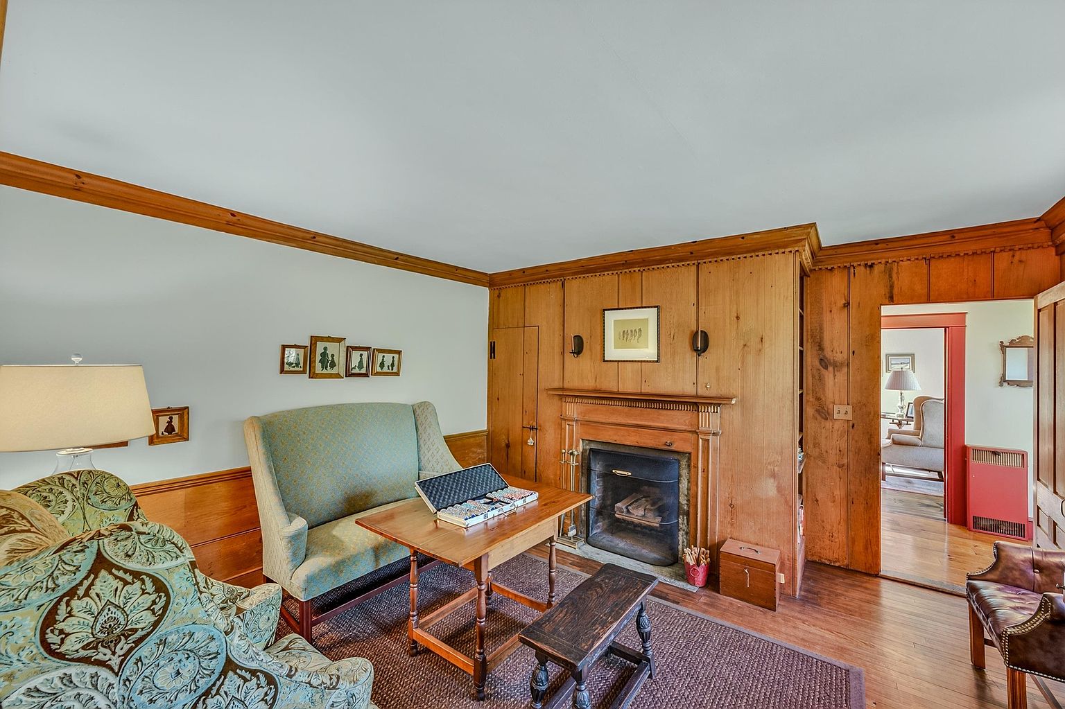 This cozy living room features warm wood-paneled walls and a traditional fireplace with a wooden mantel as the central focal point. The space is furnished with a patterned armchair, a light-colored sofa, and a wooden table set up for games, all resting on a textured area rug. A doorway leads into an adjacent room, creating an inviting and classic atmosphere with a rustic, homey aesthetic.