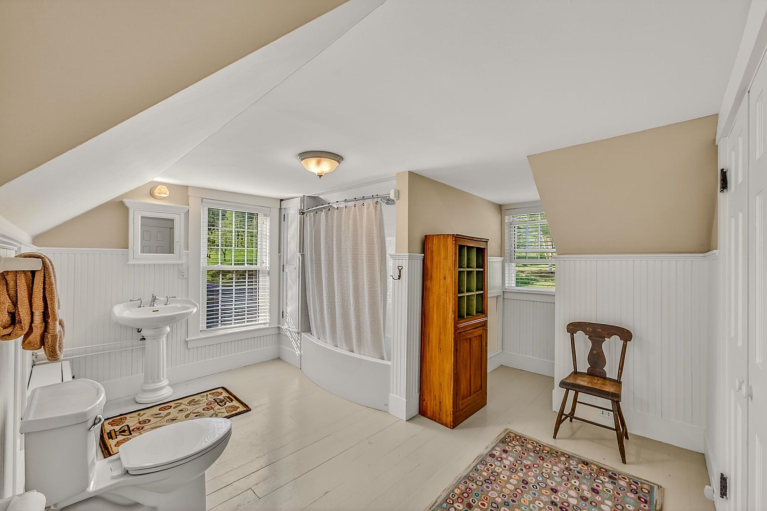 This bright, attic-level bathroom features a classic pedestal sink, a white toilet, and a shower-tub combination enclosed by a white curtain. The room is characterized by its sloped ceilings, white beadboard wainscoting, and light-colored wood flooring accented by two patterned area rugs. A tall, light wood cabinet provides storage, while a simple wooden chair adds a rustic touch to the space.