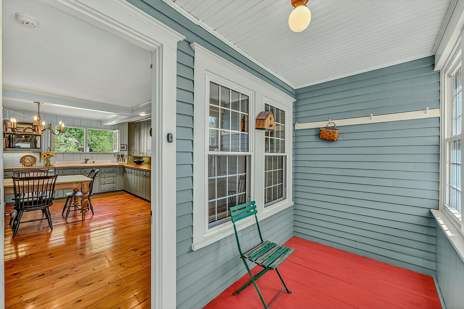 This charming enclosed porch features vibrant red painted flooring and light blue horizontal siding, creating a cozy and inviting atmosphere. A single green metal chair sits in the foreground, while a doorway leads directly into a bright kitchen with warm wood floors and classic cabinetry. The space is illuminated by a simple overhead light fixture and large windows that allow natural light to flow between the two rooms.