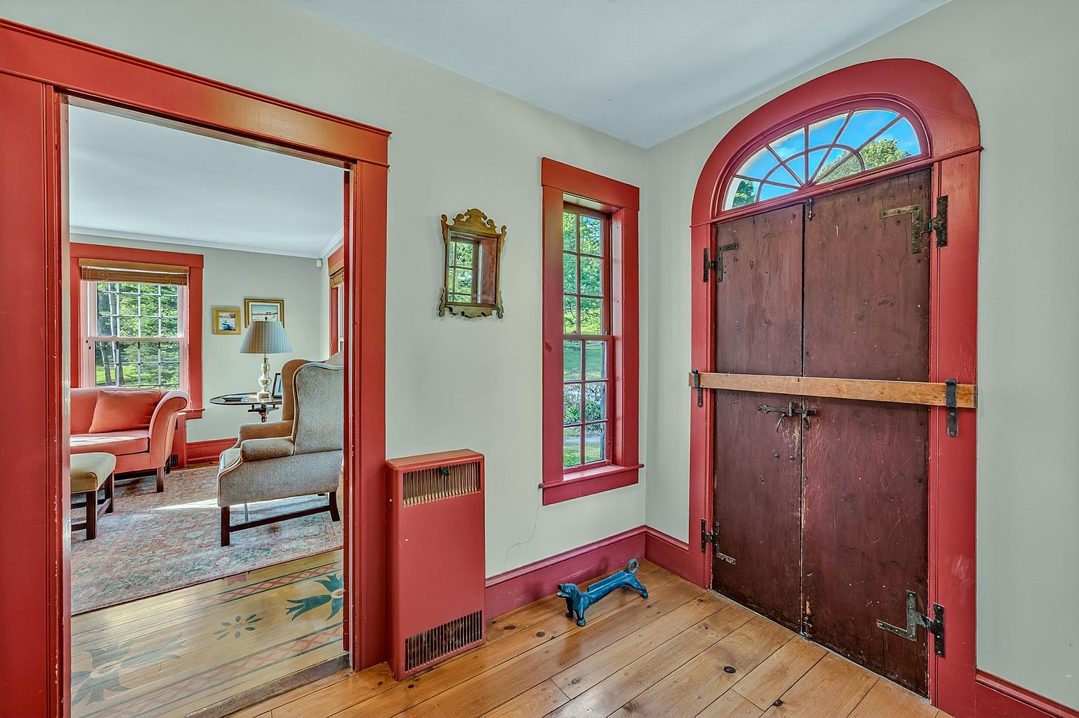 This entryway features warm, natural wood flooring and a striking double door with a decorative arched transom window, all framed by bold red trim. A view through the wide doorway reveals a cozy living area with a sofa and armchair, creating a sense of depth and flow between rooms. The space is accented by a small wall mirror and a unique blue metal boot scraper, contributing to a charming, historic aesthetic.