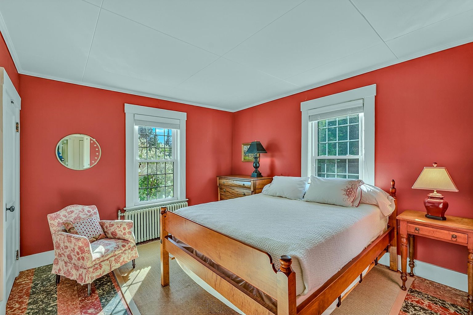 This cozy bedroom features vibrant red walls and a classic wooden bed frame centered in the room. A patterned armchair sits in the corner near a window, while a wooden side table with a lamp provides a warm, traditional aesthetic. The room is bathed in natural light from two windows, creating a bright and inviting atmosphere.