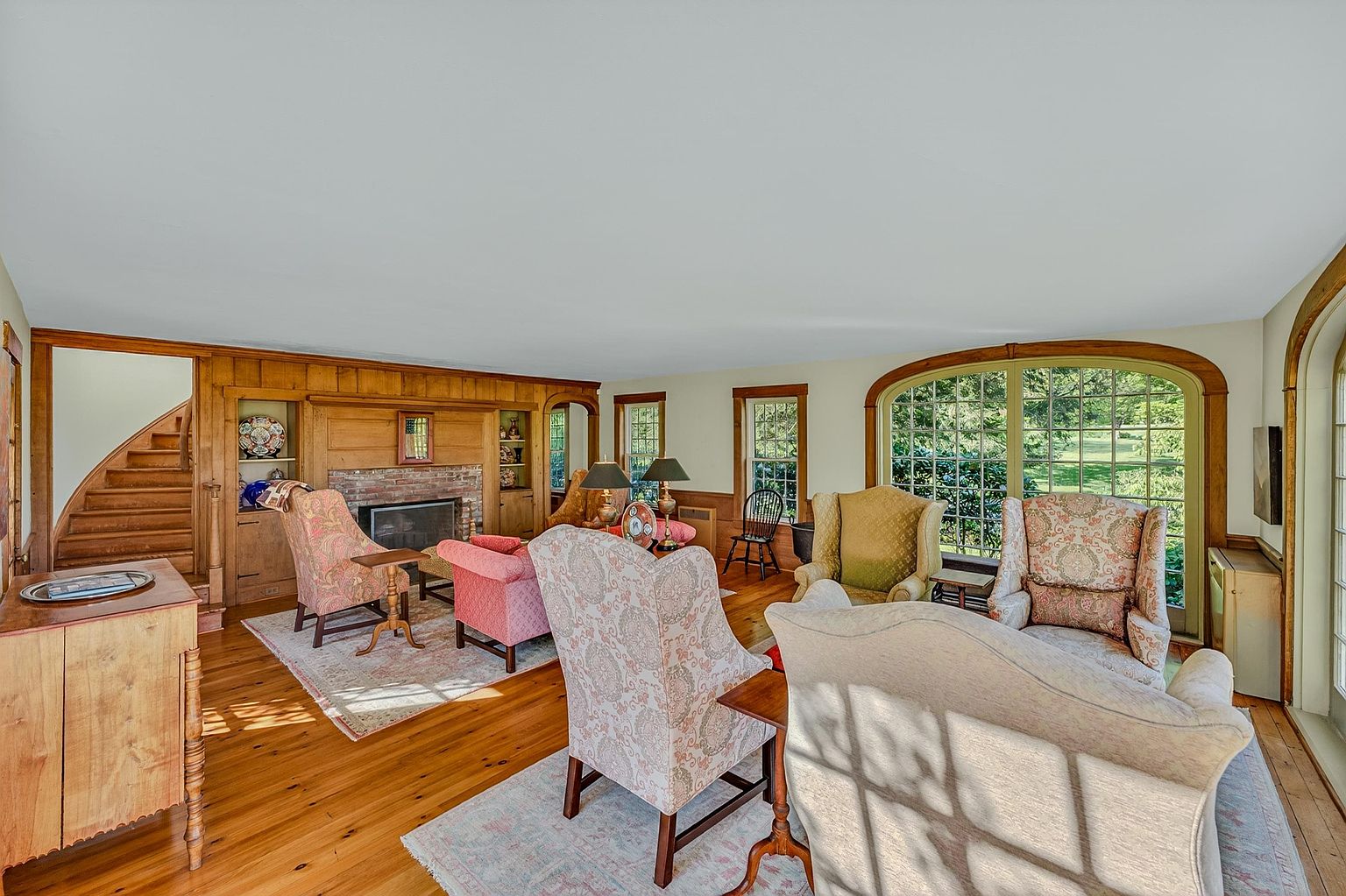 This spacious living room features warm wood paneling, hardwood floors, and a brick fireplace surround that serves as a focal point. The room is furnished with multiple patterned armchairs and a sofa, arranged to create a comfortable seating area, while large arched windows provide ample natural light and views of the outdoors. A staircase with a wooden banister is visible on the left, adding architectural interest to this traditional, inviting space.