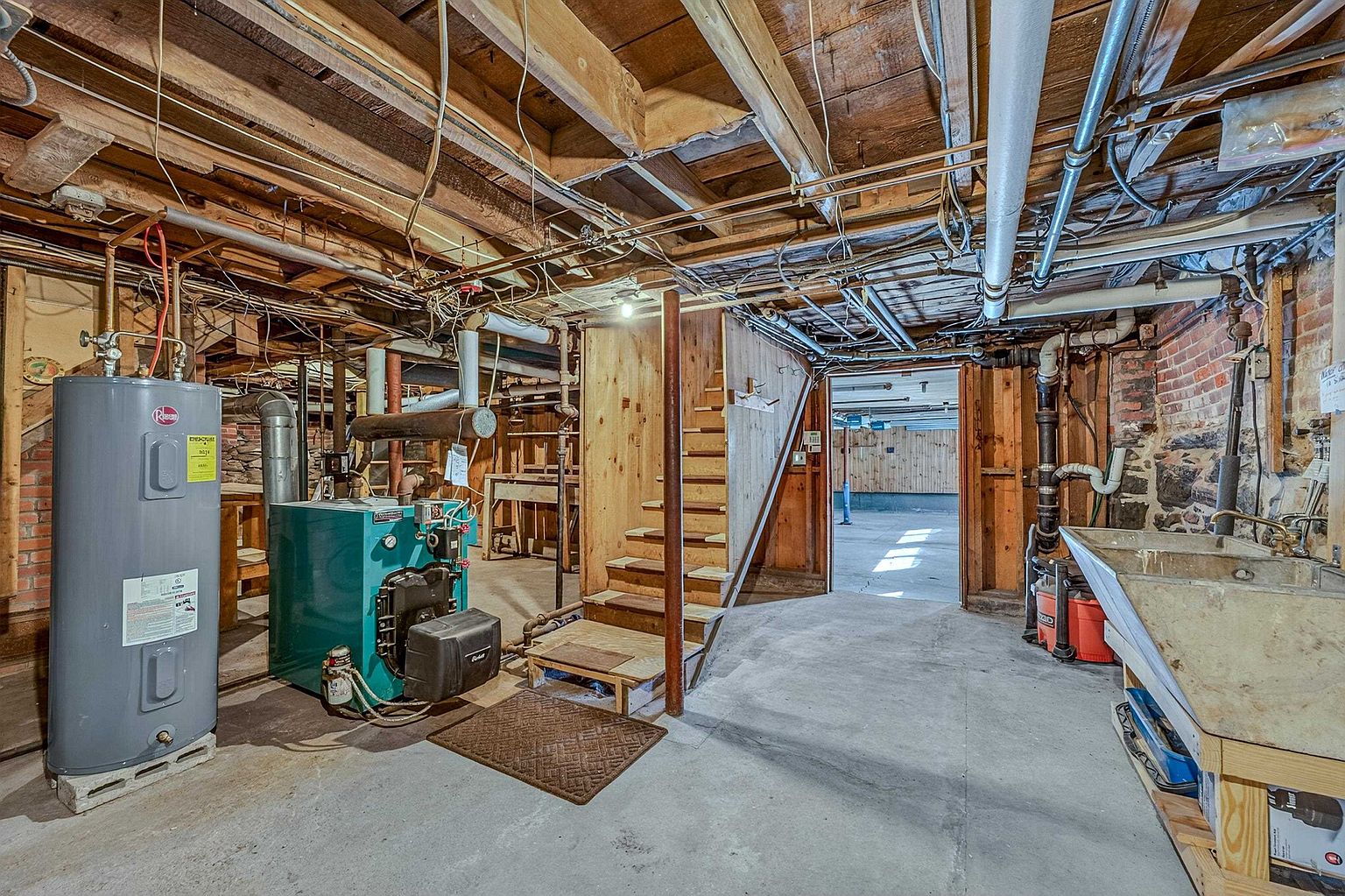 This unfinished basement features exposed wooden ceiling joists, concrete flooring, and a mix of stone and wood-paneled walls. The space includes a utility area with a water heater and a boiler, a set of wooden stairs leading to an adjacent room, and a utility sink. The overall impression is that of a functional, utilitarian storage or mechanical space with a rustic, industrial aesthetic.