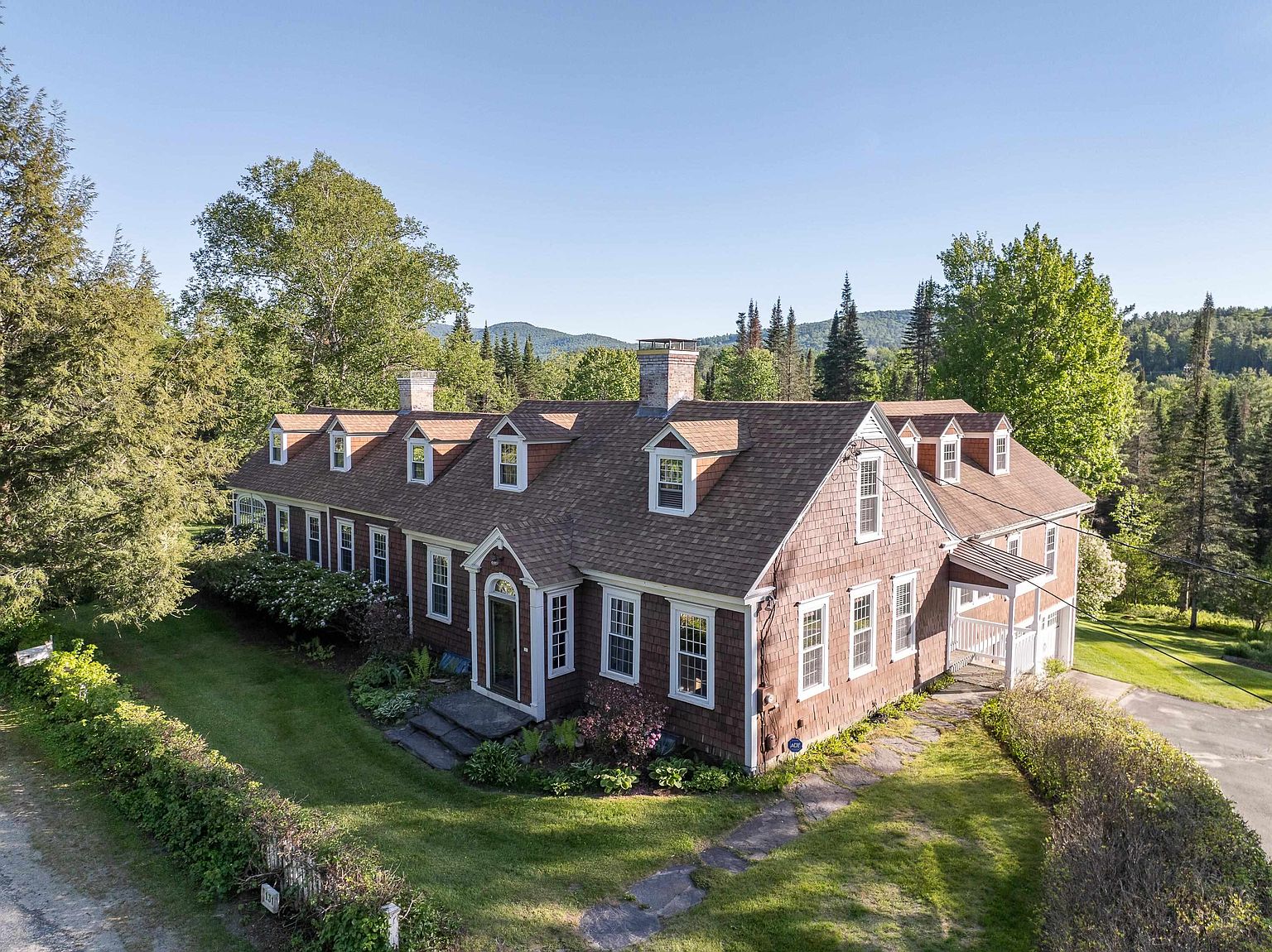 This elevated aerial view captures a charming, traditional-style home with dark brown shingle siding and a multi-gabled roofline featuring several dormer windows. The property is nestled in a lush, green landscape with mature trees and rolling hills in the background, creating a serene and private atmosphere. A stone pathway leads to the front entrance, and the well-maintained lawn and surrounding foliage enhance the home's classic, rustic appeal.