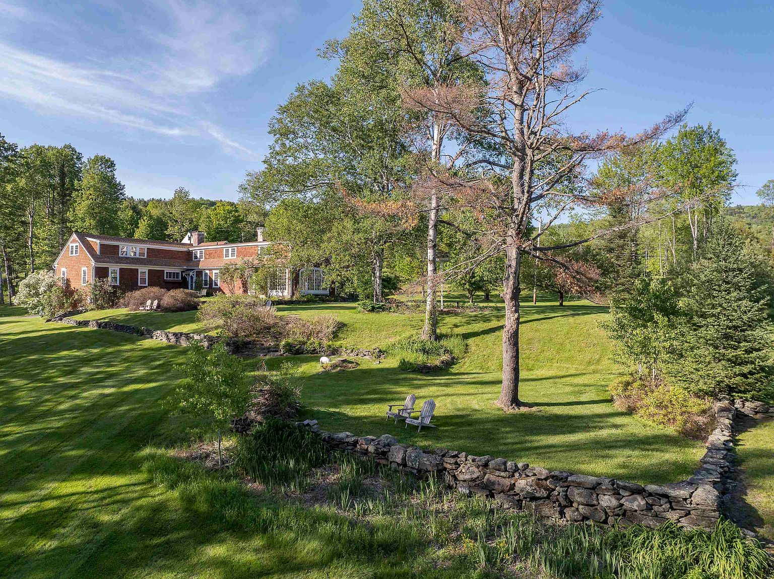 This expansive property features a traditional shingled home nestled into a lush, rolling landscape. The foreground showcases a rustic stone wall and a manicured lawn with two Adirondack chairs positioned under a large, mature tree. The scene is captured from a slightly elevated perspective, highlighting the serene, park-like setting and the home's integration with the surrounding woodland.