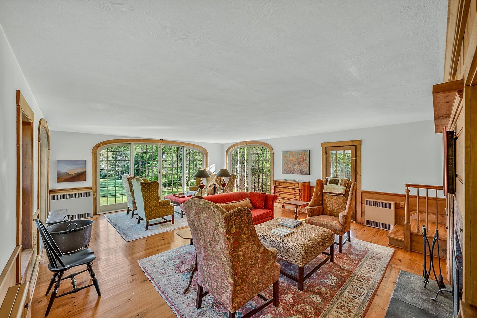 This spacious living room features warm wood flooring and a traditional aesthetic with multiple seating areas, including a red sofa and several patterned armchairs. Large, arched windows allow for abundant natural light and provide views of the lush greenery outside. A stone hearth sits to the right, anchoring the room, while the overall atmosphere is inviting and classic.