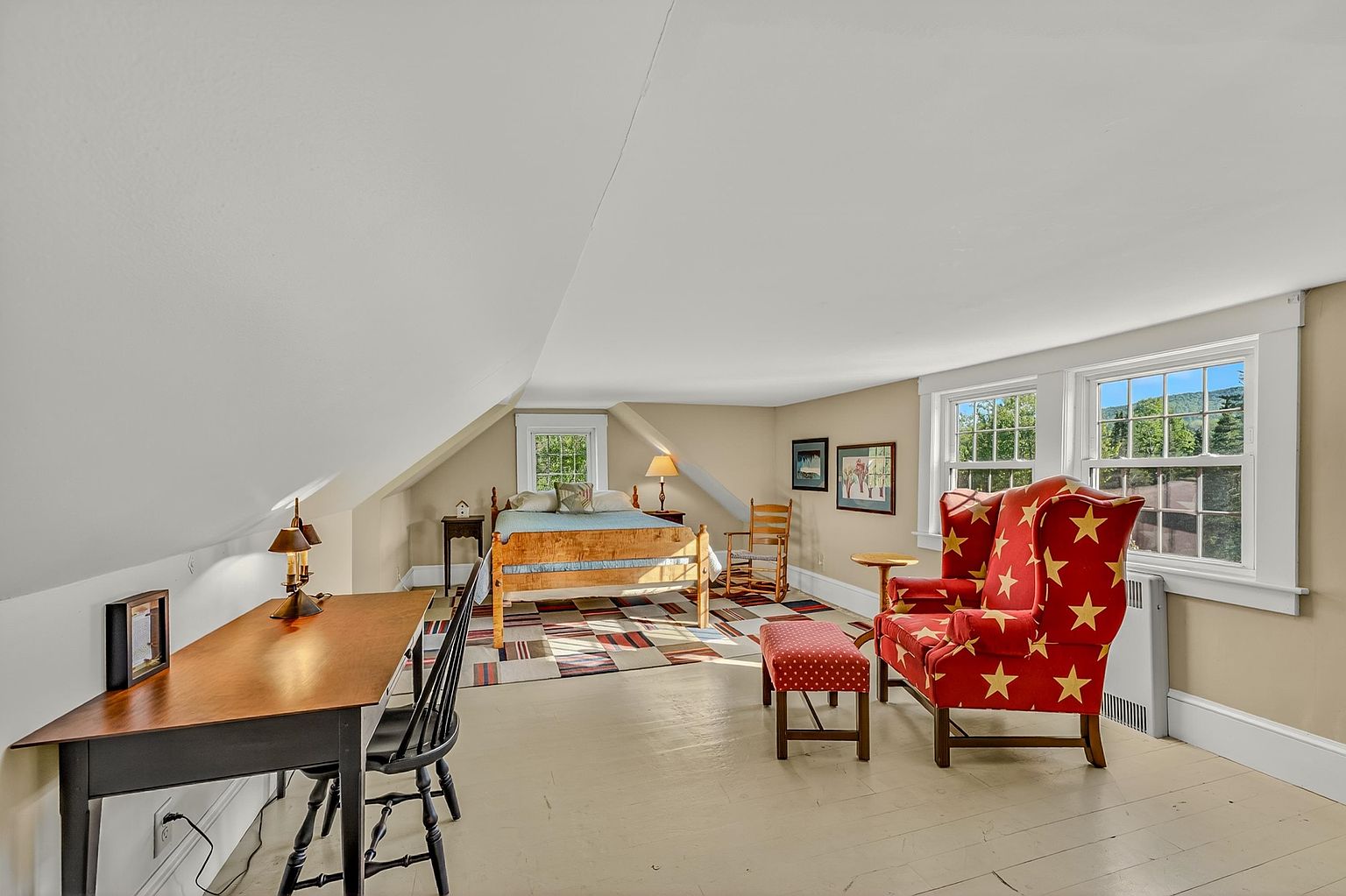 This spacious attic bedroom features a sloped ceiling and light-colored wood flooring, creating a cozy and inviting atmosphere. The room is furnished with a wooden bed frame, a vibrant red armchair with star patterns, and a dedicated workspace with a desk and chair. Multiple windows allow natural light to fill the space, offering views of the surrounding greenery.