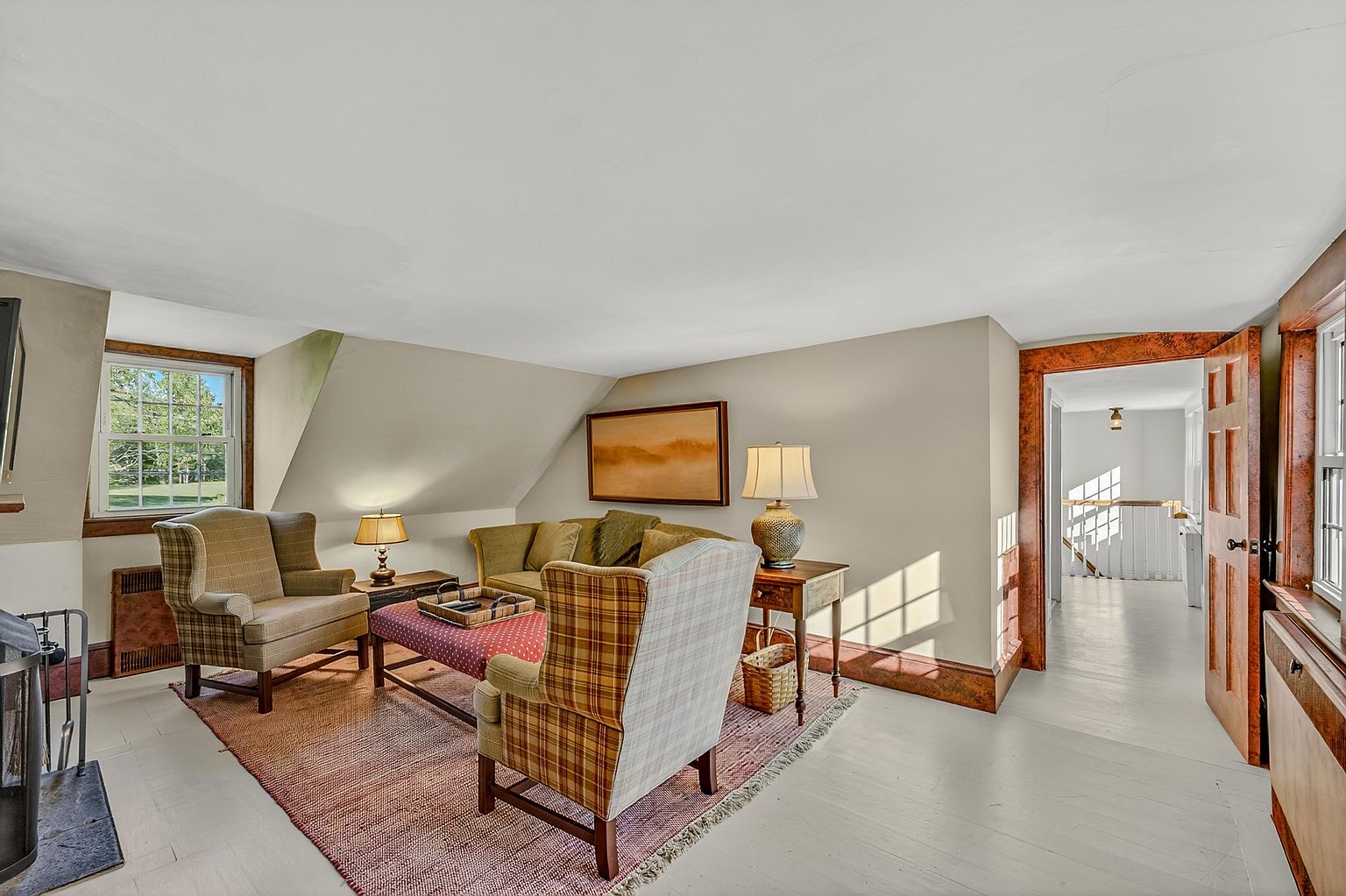 This cozy living room features a sloped ceiling and a warm, traditional aesthetic with a mix of plaid and solid-colored upholstered seating. A central seating area is anchored by a textured rug, while a wooden side table with a lamp and a framed landscape painting add character to the space. The room flows into an adjacent hallway, with natural light streaming in from windows on both sides, creating an inviting and comfortable atmosphere.