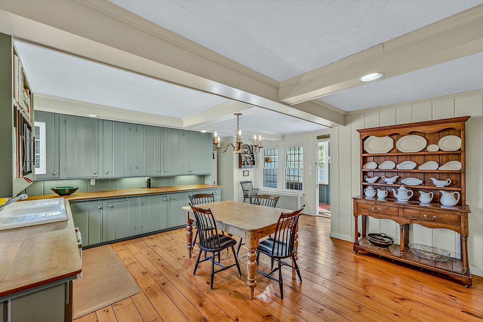 This spacious, rustic-inspired kitchen features sage green cabinetry paired with warm wood countertops and wide-plank wood flooring. A central dining area with a wooden table and black spindle chairs sits beneath a classic chandelier, while a large hutch displays a collection of white ceramic dishware. The room is bright and inviting, with a seamless flow between the cooking area and the dining space, accented by exposed ceiling beams.