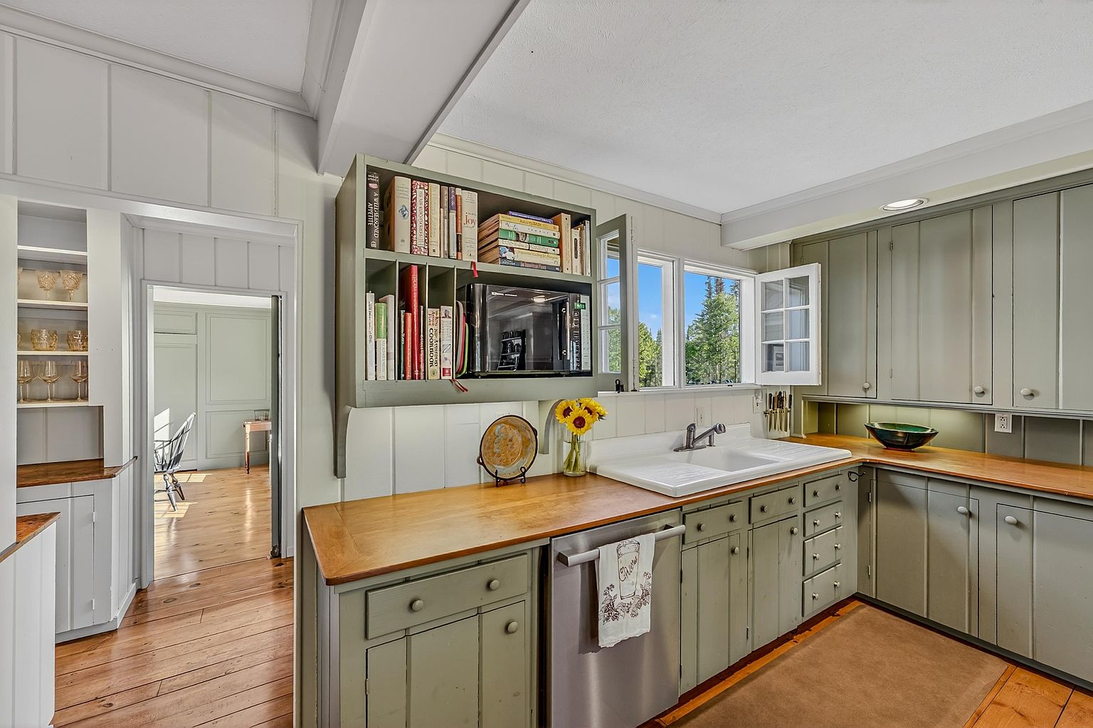 This charming, rustic-inspired kitchen features sage green cabinetry paired with warm wood countertops and a classic white farmhouse sink. A built-in shelving unit above the counter provides convenient storage for cookbooks and a microwave, while a window above the sink brings in natural light. The space flows seamlessly into an adjacent room, showcasing hardwood flooring and a cozy, traditional aesthetic.