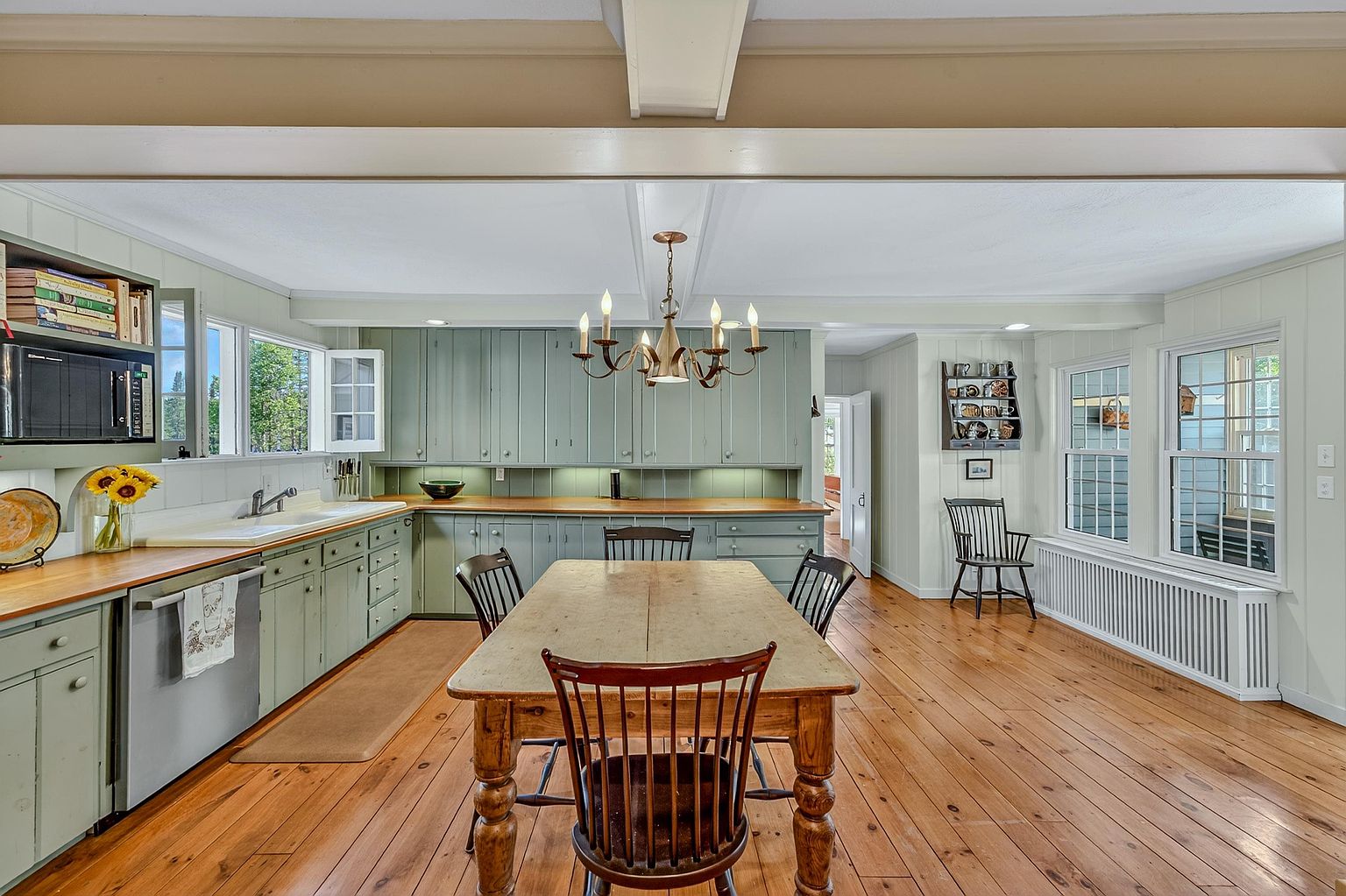 This charming, rustic-inspired kitchen features sage green cabinetry paired with warm wood countertops and wide-plank wood flooring. A central wooden dining table serves as a focal point under a classic chandelier, while large windows allow natural light to fill the space. The room maintains a cozy, traditional aesthetic with open shelving and a functional layout that flows into an adjacent living area.