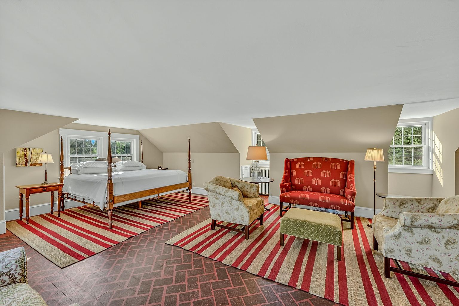 This spacious primary bedroom features a classic four-poster bed and a cozy seating area arranged on a large, striped area rug. The room is characterized by its unique sloped ceilings, brick-patterned flooring, and multiple windows that bring in natural light. The decor combines traditional furniture with a warm, inviting color palette, creating a comfortable and expansive retreat.