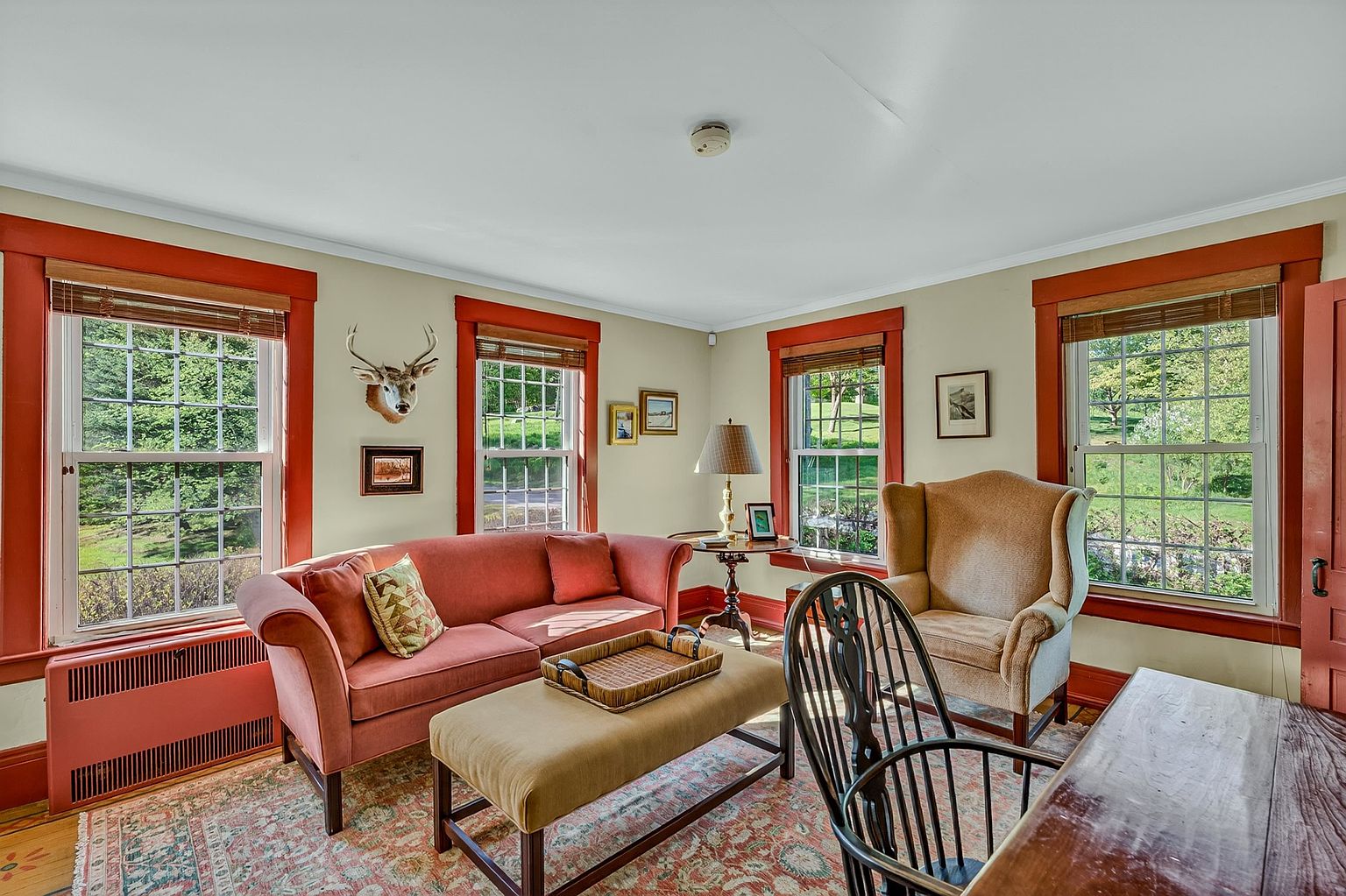 This cozy living room features a warm, inviting atmosphere with a coral-toned sofa, a matching ottoman with a woven tray, and a comfortable wingback chair. The space is framed by large windows with distinctive reddish-orange trim that look out onto a lush, green landscape. A patterned area rug anchors the room, while a deer mount and framed artwork add a touch of traditional, rustic character to the walls.