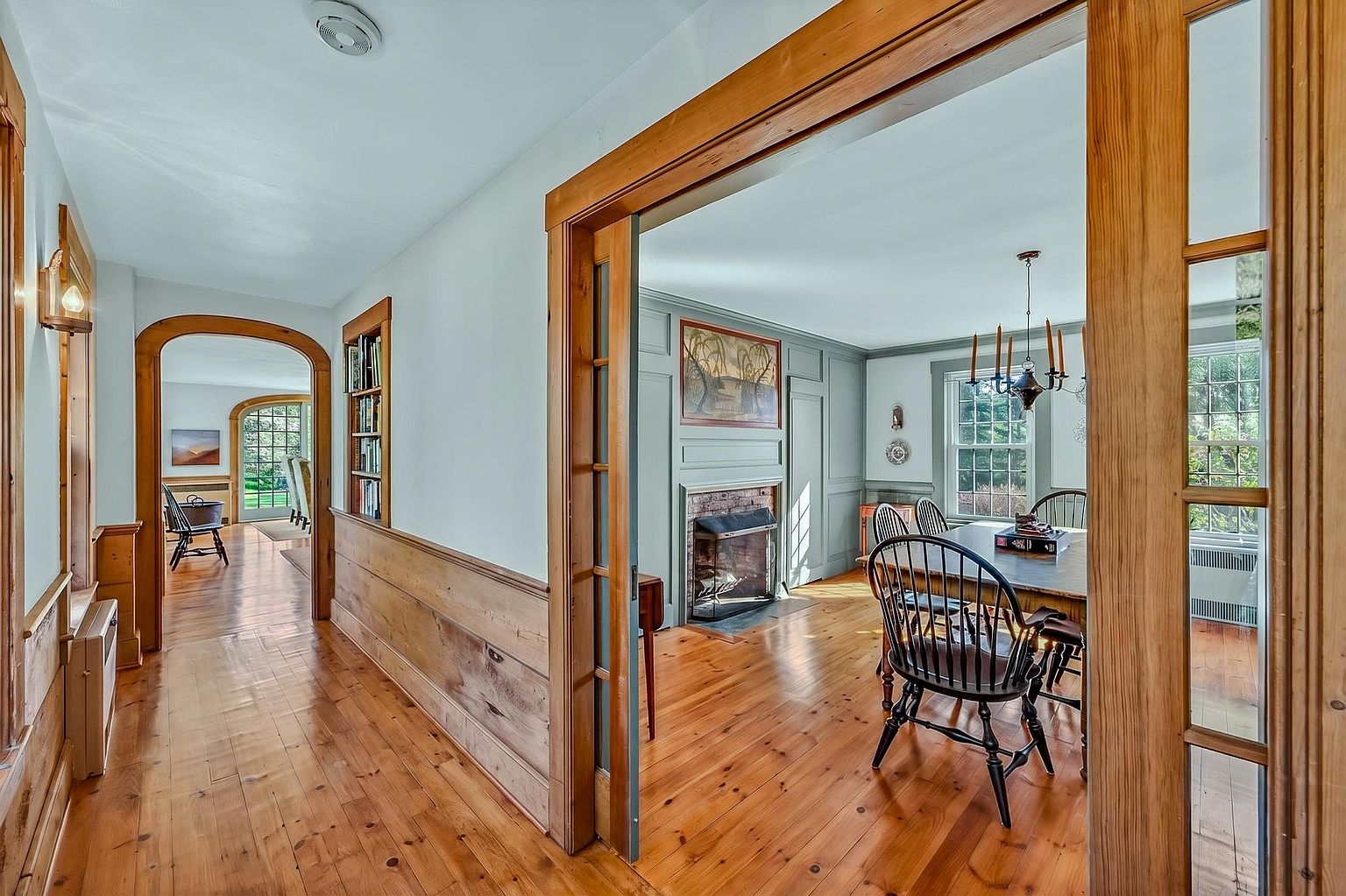 This perspective captures a transition space featuring warm wood flooring and wainscoting that flows from a hallway into a formal dining room. The dining area is anchored by a brick fireplace with a wood mantel and a classic chandelier, while the hallway includes a built-in bookshelf and an arched doorway leading to another room. The home exhibits a traditional, character-filled aesthetic with natural wood trim and soft-toned walls.