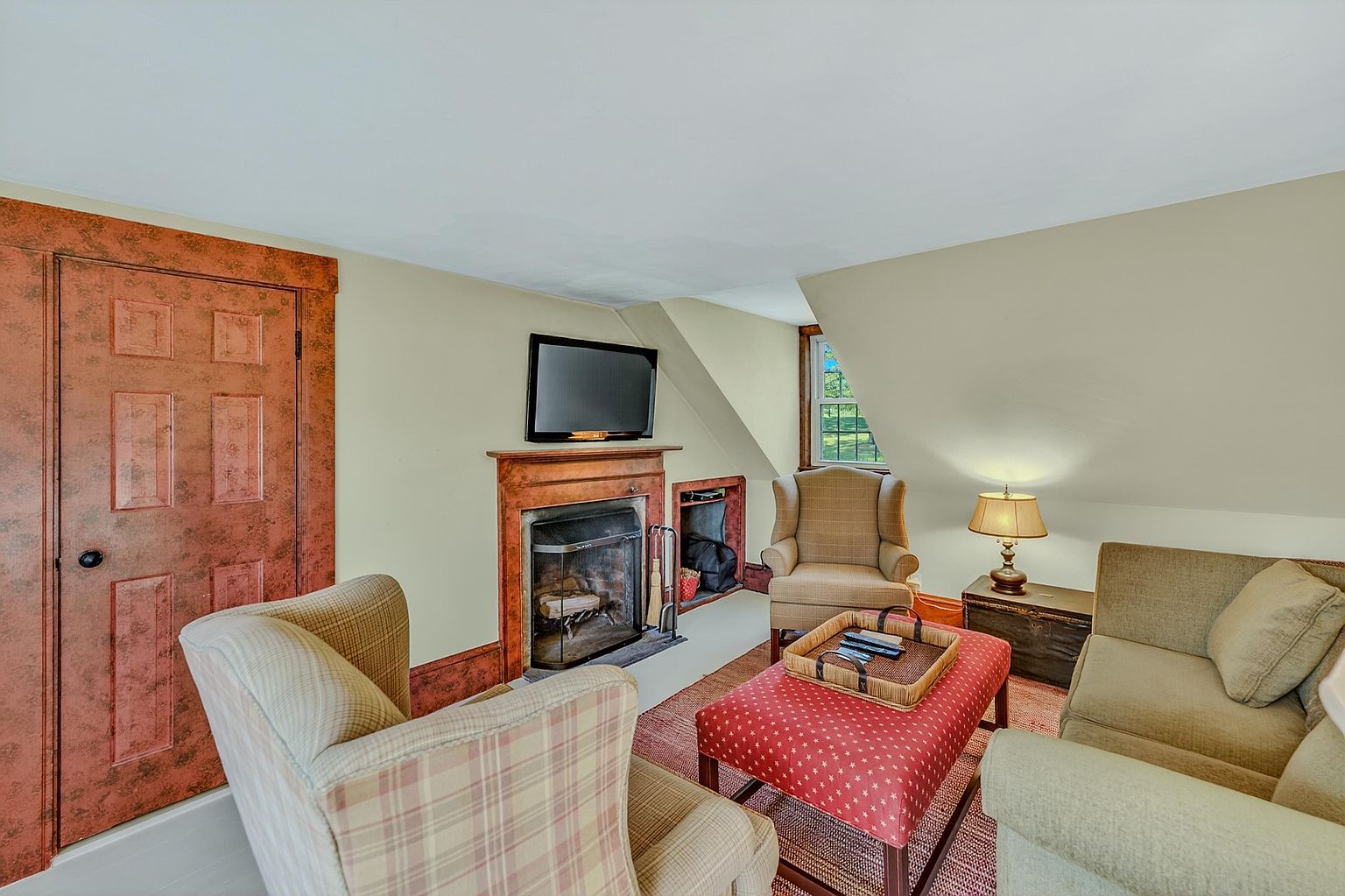 This cozy living room features a warm, traditional aesthetic with a prominent fireplace as the focal point, topped by a wall-mounted television. The space is furnished with a patterned armchair, a matching red ottoman with a serving tray, and a neutral-toned sofa, all arranged on a textured rug. Sloped ceilings and a window overlooking the outdoors add character to this intimate, comfortable seating area.