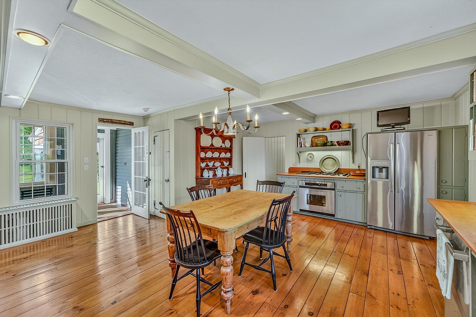 This rustic-inspired kitchen and dining area features warm wood flooring and light-toned paneled walls, creating a cozy, traditional atmosphere. A central wooden dining table is surrounded by black spindle-back chairs, positioned beneath a classic chandelier and a large wooden hutch. The kitchen workspace includes stainless steel appliances, a built-in cooktop, and open shelving, all set against a backdrop of soft-colored cabinetry.