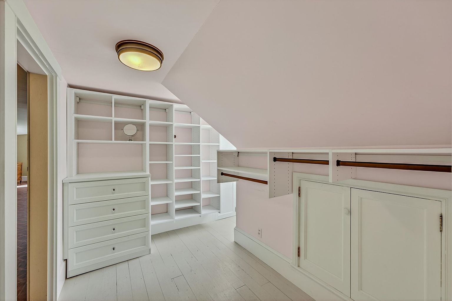 This walk-in closet features a sloped ceiling and light-colored walls, creating a bright and airy atmosphere. The space is efficiently organized with built-in white shelving, a four-drawer dresser, and hanging rods tucked beneath the eaves. The light-toned wood flooring and simple flush-mount ceiling light complete the clean, functional design.