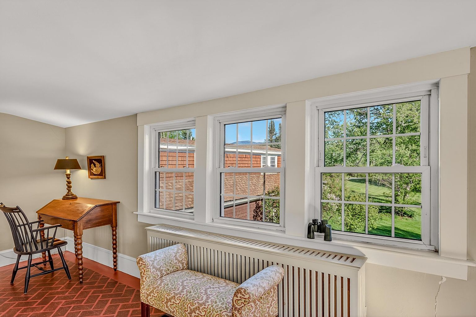 This bright office space features a classic wooden writing desk paired with a spindle-back chair, set against a backdrop of patterned tile flooring. A long radiator cover sits beneath three large windows that offer a view of the outdoors, while a small framed artwork hangs on the wall above the desk. The room has a traditional, cozy atmosphere with neutral wall tones and ample natural light.