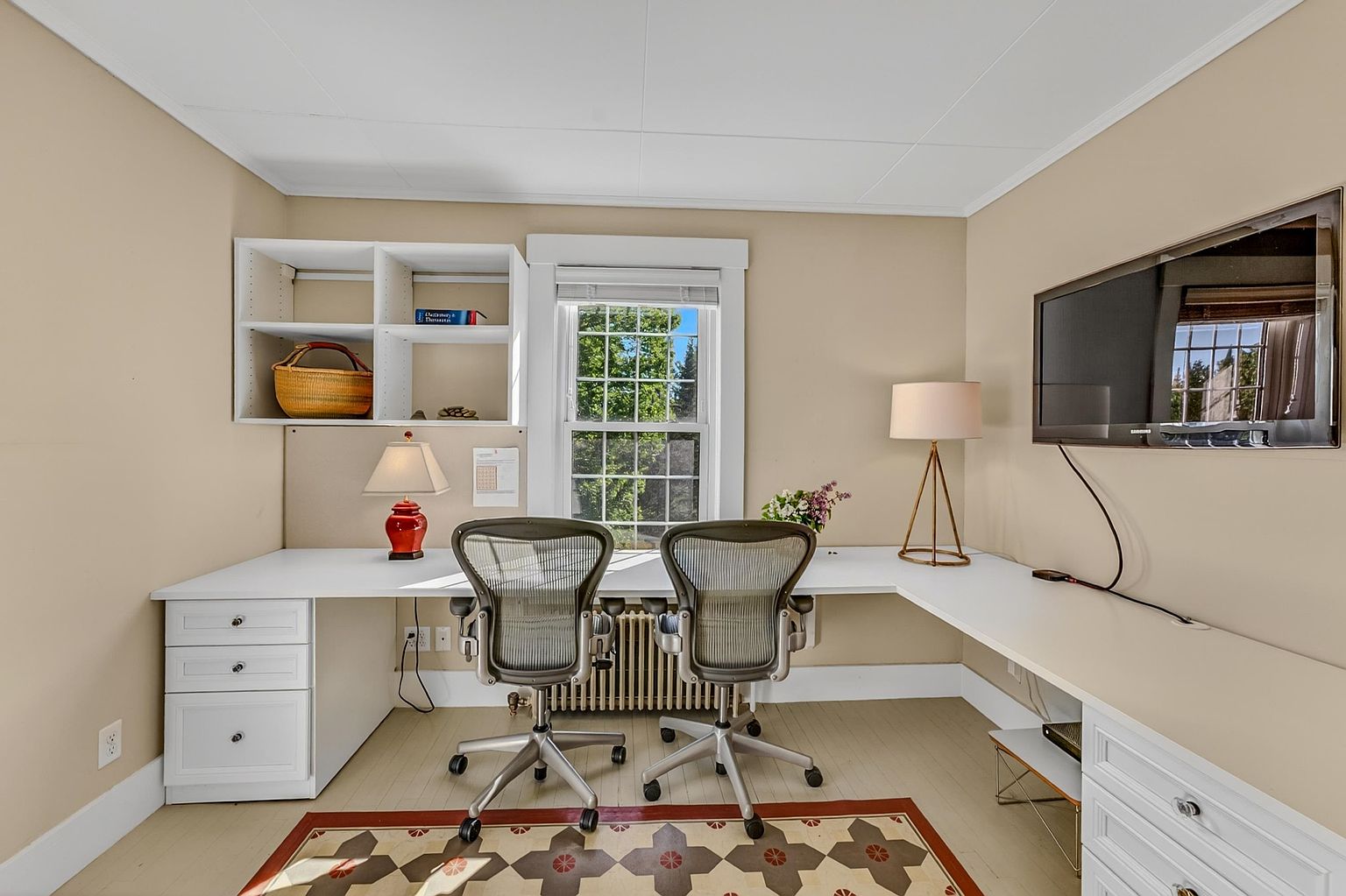 This home office features a spacious L-shaped desk configuration with white cabinetry and ample workspace for two. The room is accented by a patterned area rug, a central window providing natural light, and built-in shelving above the desk. A wall-mounted television and two ergonomic office chairs complete this functional and well-lit workspace.