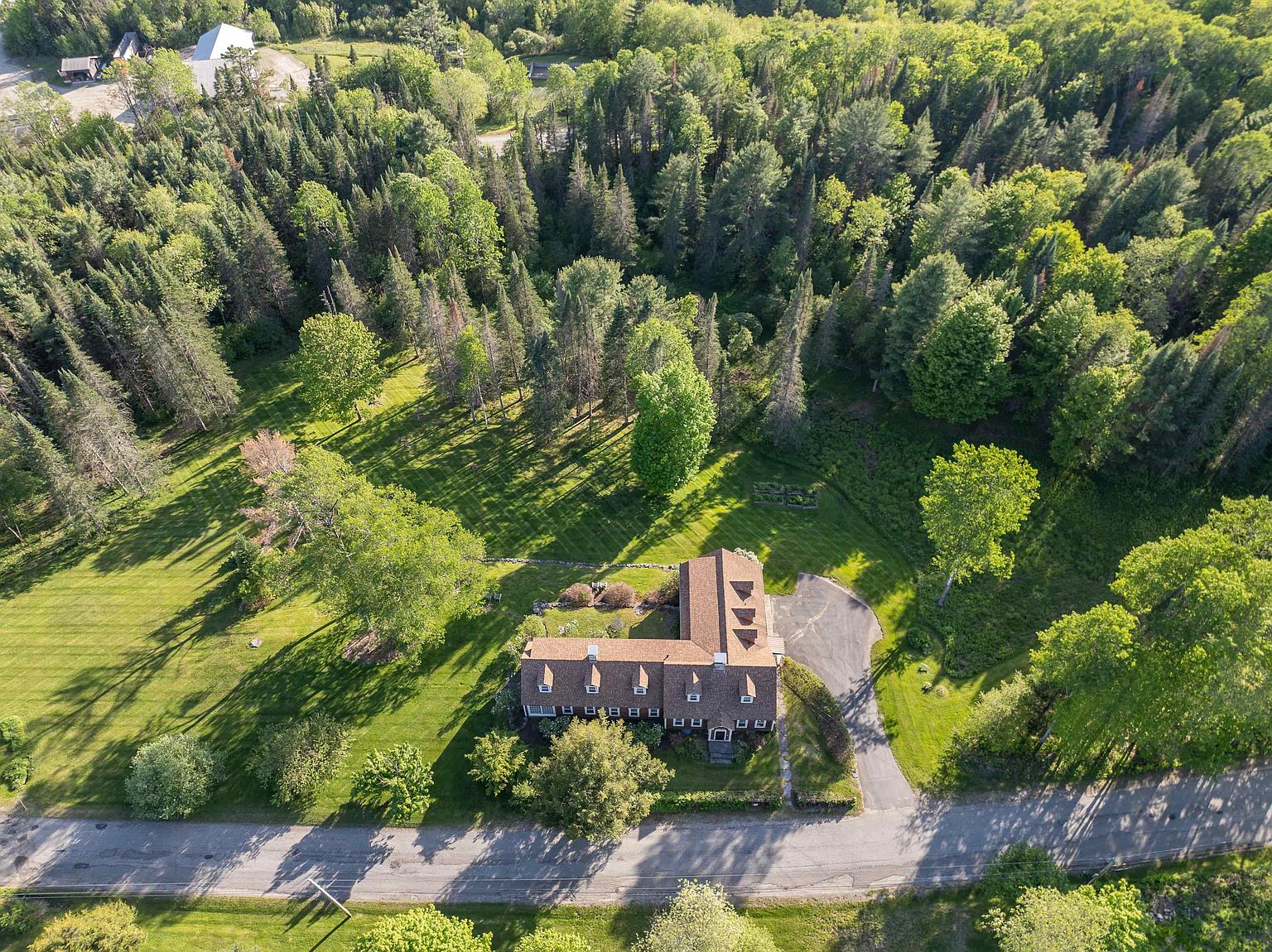 This high-angle aerial view captures an L-shaped, multi-story residence nestled within a lush, wooded landscape. The home features a classic brown shingled roof with multiple dormer windows and a paved driveway leading to the side entrance. The property is surrounded by expansive green lawns and dense forest, creating a serene and private setting.