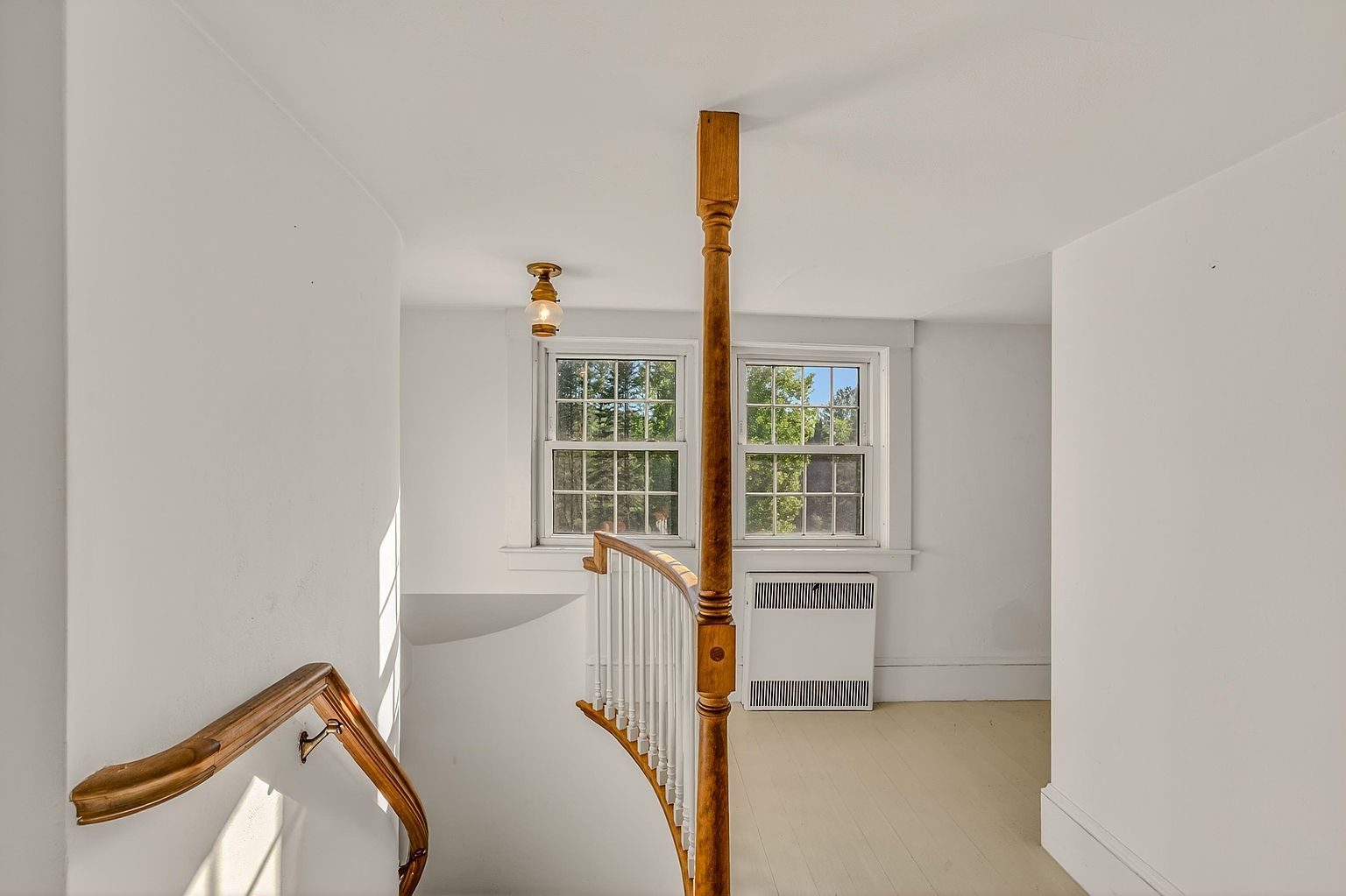 This image captures a view from the top of a staircase looking toward a landing with two windows. The space features white walls, light-toned flooring, and a prominent wooden banister with a decorative newel post. A white radiator sits beneath the windows, and a simple light fixture hangs from the ceiling, creating a bright and traditional atmosphere.