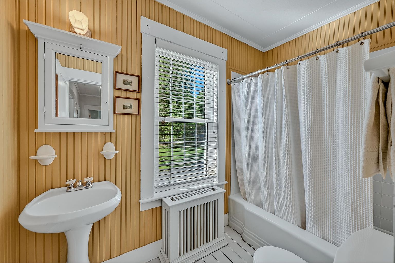 This bathroom features a classic pedestal sink, a white tub with a shower curtain, and a medicine cabinet with integrated lighting. The walls are finished with vertical wood paneling in a warm yellow tone, complemented by white trim and white painted wood flooring. A window with white blinds provides natural light, and a radiator cover sits neatly beneath it, creating a cozy and traditional aesthetic.