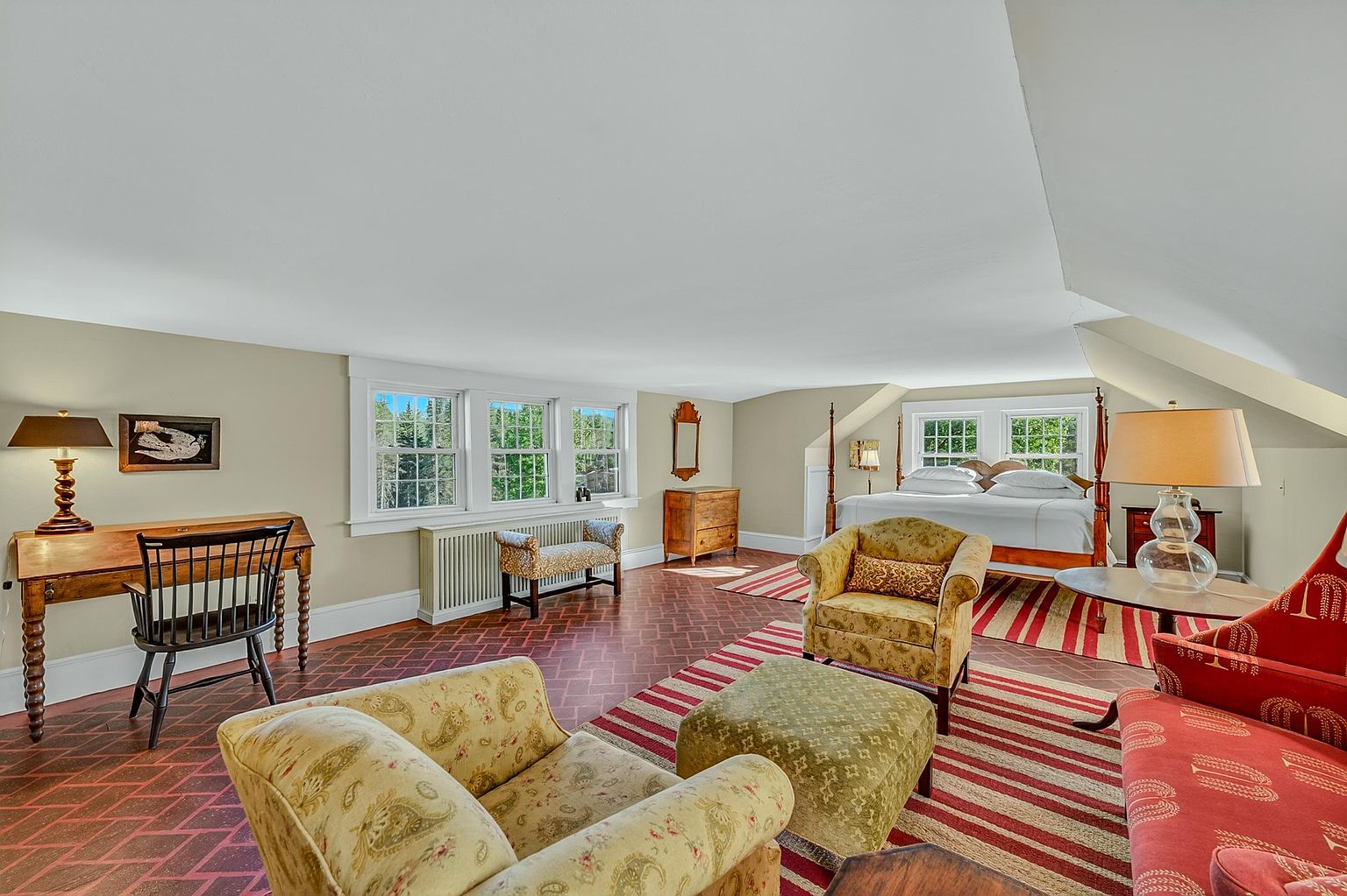 This spacious primary bedroom features a unique layout with a sloped ceiling and a distinctive patterned tile floor. The room is furnished with a four-poster bed, a cozy seating area with patterned armchairs and an ottoman, and a dedicated desk space near the windows. The warm, neutral wall color and classic furniture pieces create a comfortable and inviting atmosphere.
