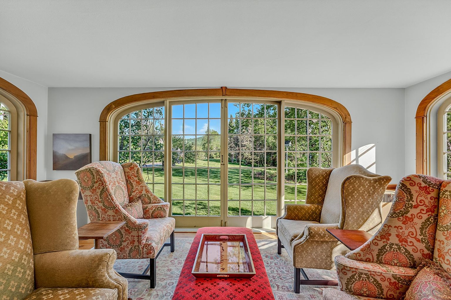This inviting living room features a symmetrical arrangement of four patterned wingback chairs centered around a long, red upholstered ottoman with a decorative tray. The space is anchored by a large, arched window wall that provides a clear view of the lush green lawn and trees outside. The room has a classic, traditional aesthetic with light-colored walls and a patterned area rug, creating a bright and comfortable atmosphere.