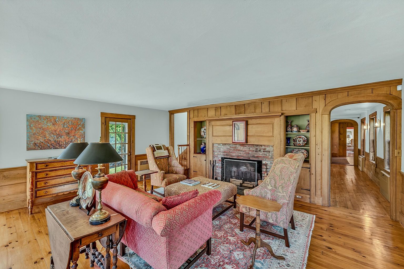 This cozy living room features warm wood paneling and a brick fireplace surround, creating a traditional and inviting atmosphere. The space is furnished with a red sofa, patterned armchairs, and a central ottoman, all arranged on a decorative area rug atop polished wood floors. A wide arched doorway leads to an adjacent hallway, while a wooden dresser and table lamps add functional charm to the room.
