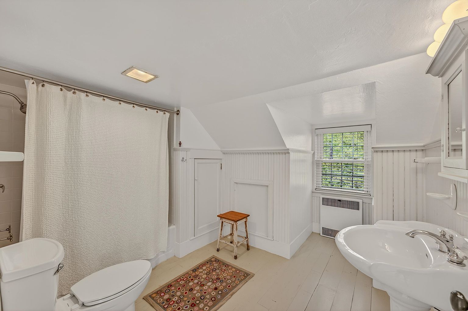 This bright, vintage-inspired bathroom features white beadboard wainscoting and a sloped ceiling that adds architectural character. The space includes a pedestal sink, a toilet, and a shower-tub combination with a white curtain, all set against light-toned wood flooring. A small wooden stool and a patterned rug provide a cozy, rustic touch, while a window with blinds allows natural light to fill the room.
