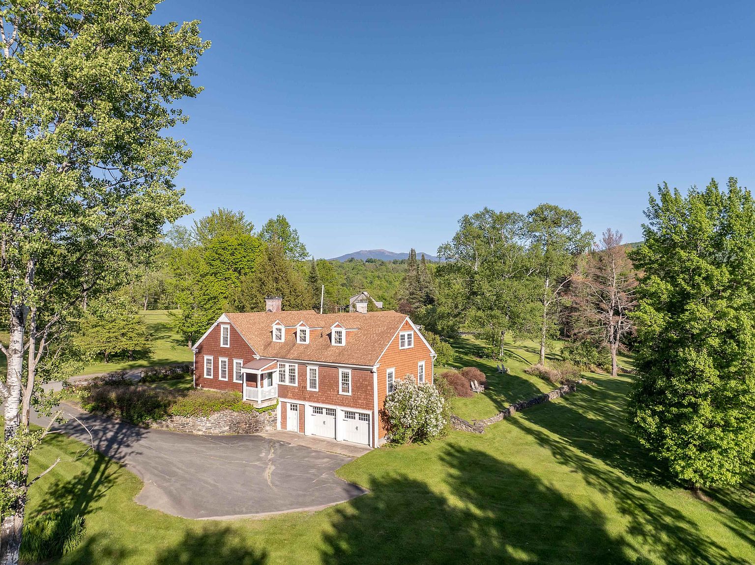 This elevated aerial perspective captures a charming, shingle-sided home nestled within a lush, expansive landscape. The property features a multi-car garage, a stone retaining wall, and a paved driveway, all set against a backdrop of rolling green hills and distant mountains. The scene conveys a serene, rural atmosphere, highlighting the home's integration into its natural, wooded surroundings.