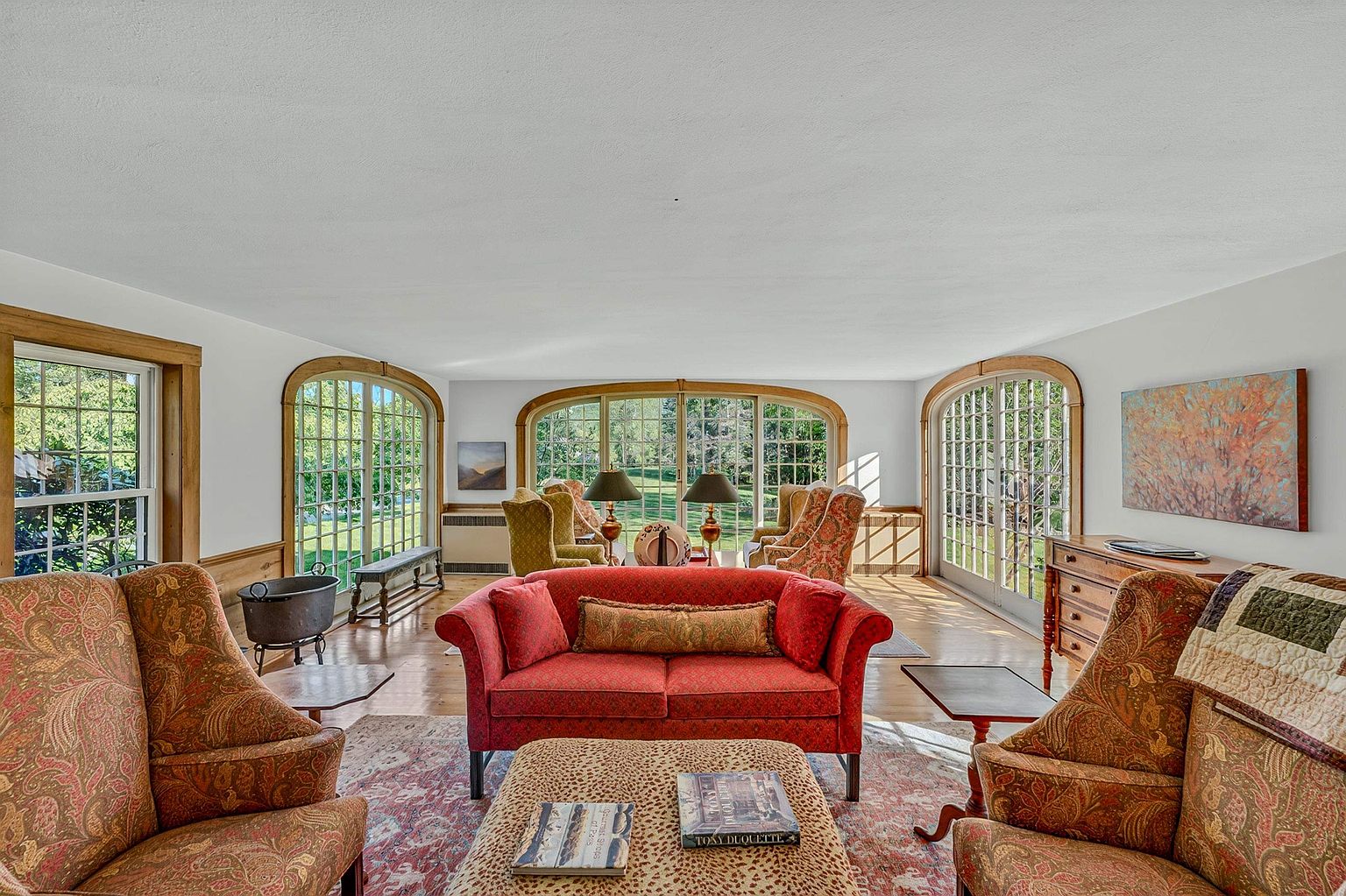 This spacious living room features a traditional design with a vibrant red sofa as the central focal point, complemented by patterned armchairs and a leopard-print ottoman. The room is framed by large, arched windows that provide abundant natural light and views of the surrounding greenery. The space is anchored by a large, ornate area rug and features a wooden dresser and side tables, creating a warm and inviting atmosphere.