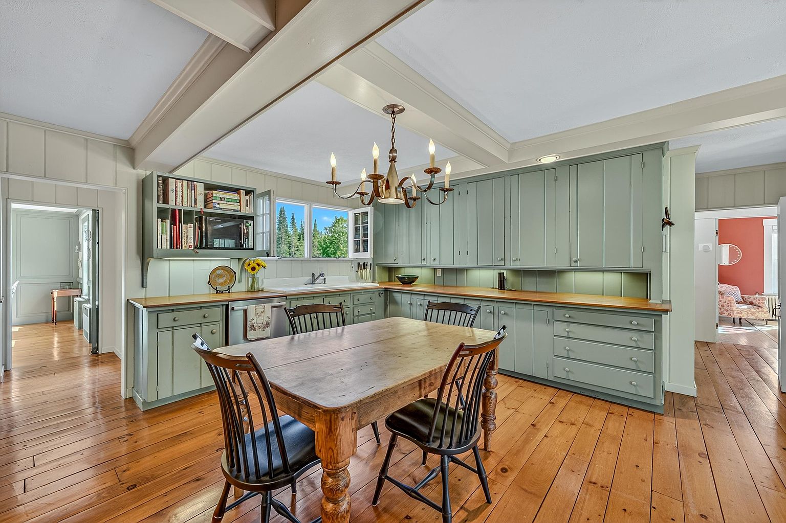 This charming, rustic-inspired kitchen features sage green cabinetry paired with warm wood countertops and wide-plank hardwood flooring. A central wooden dining table serves as a focal point under a classic chandelier, while open shelving and a farmhouse-style sink enhance the cozy, traditional aesthetic. The perspective is a wide, eye-level shot that captures the functional layout and the inviting, warm atmosphere of the space.